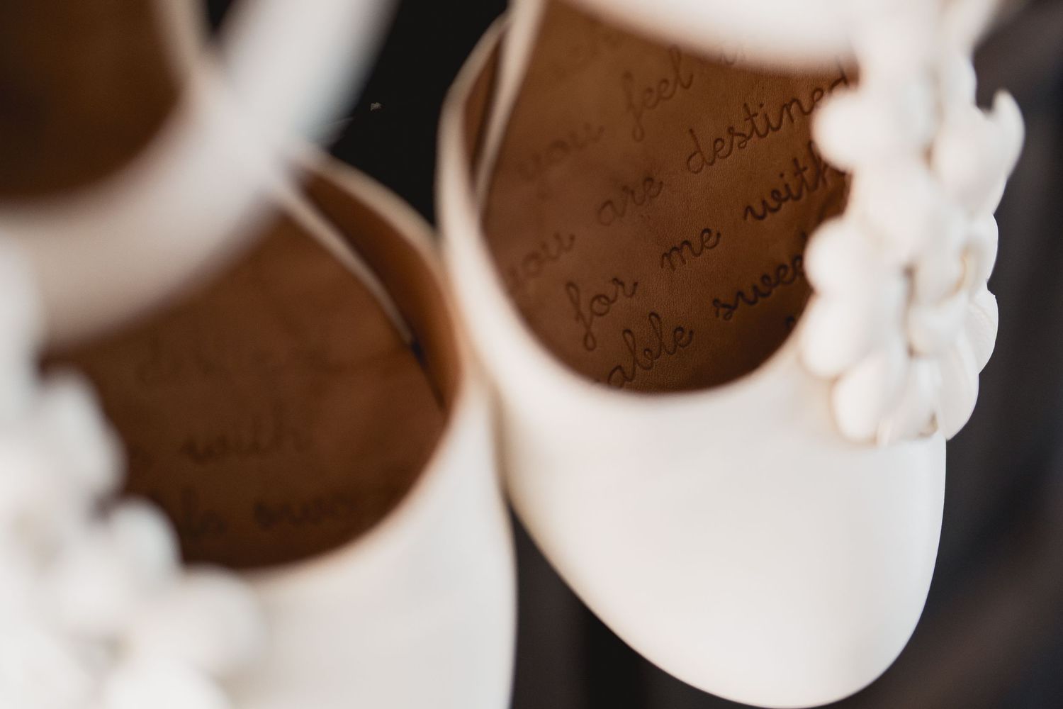 Delicate white baby shoes with floral details shown in soft focus against dark background.