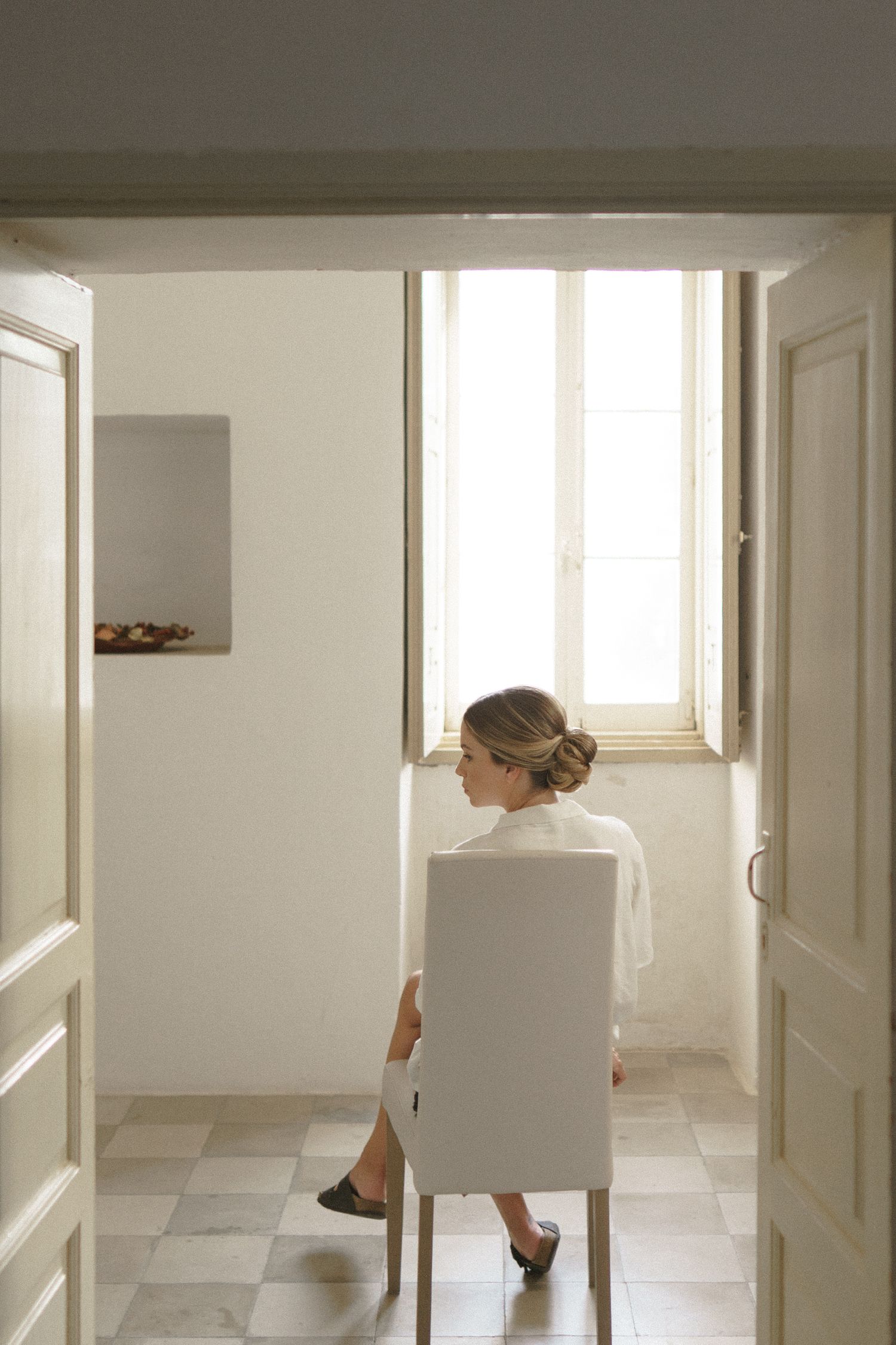 Figure in white sits alone in a bright minimalist room with checkered floor tiles near a window.
