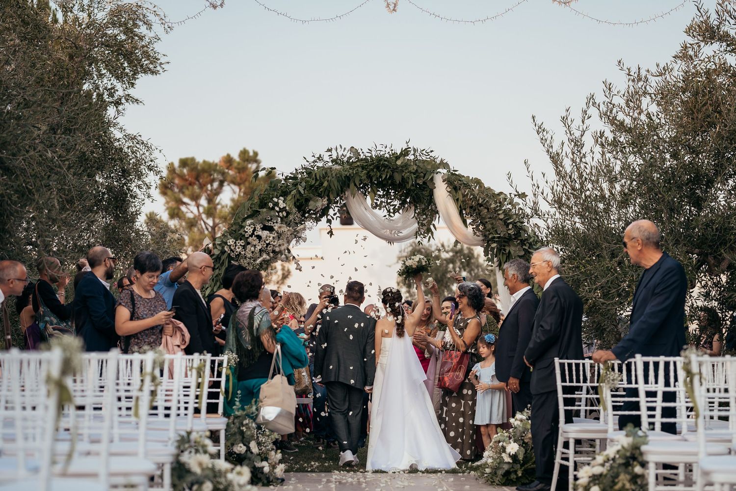 Guests throw confetti over the newlyweds during an outdoor wedding ceremony with white chairs.