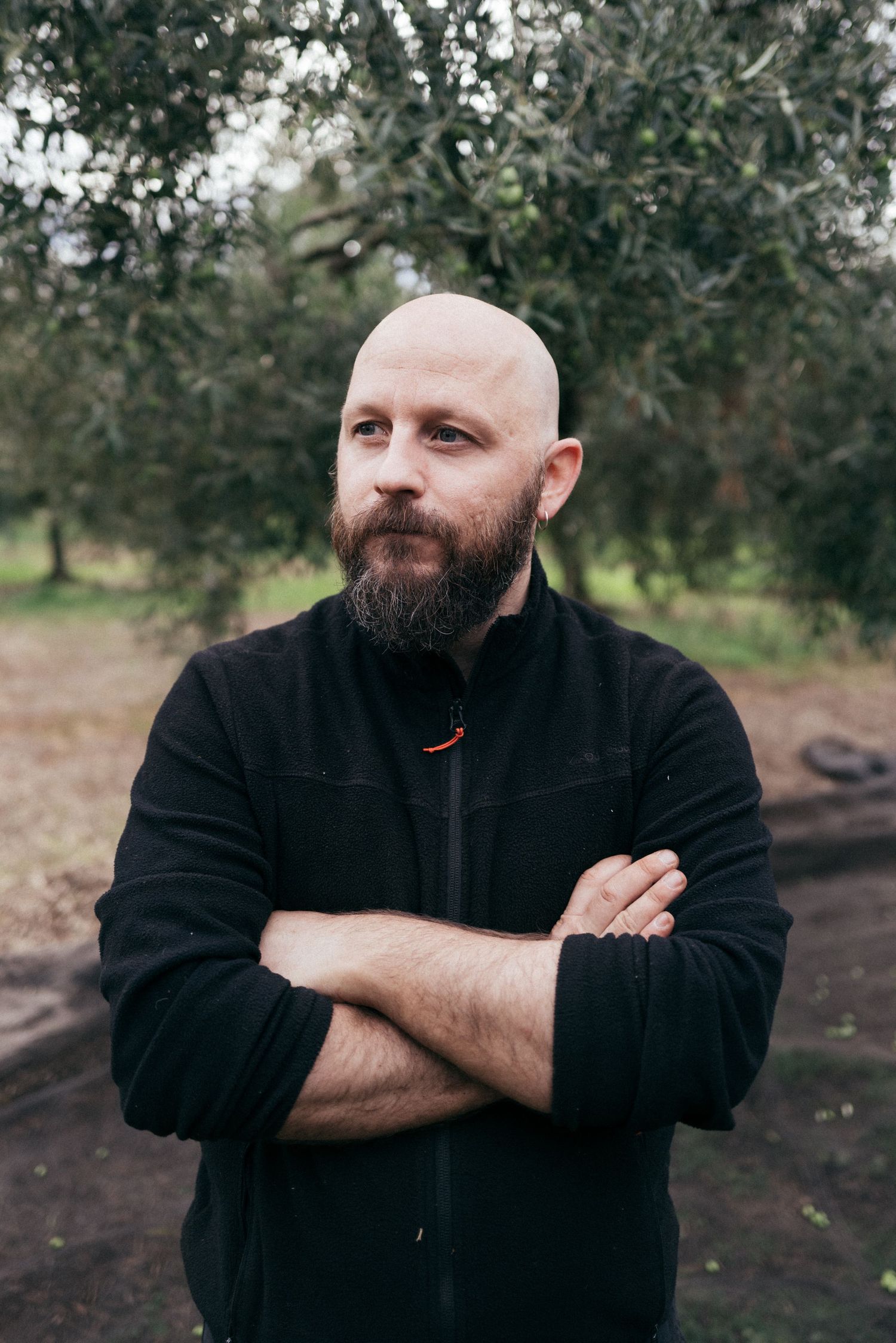 Person in black jacket with crossed arms stands among olive trees.