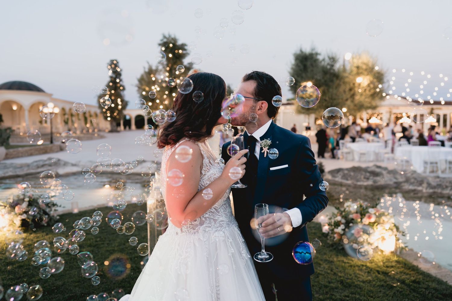 Couple sharing champagne and laughter at outdoor wedding reception with string lights.