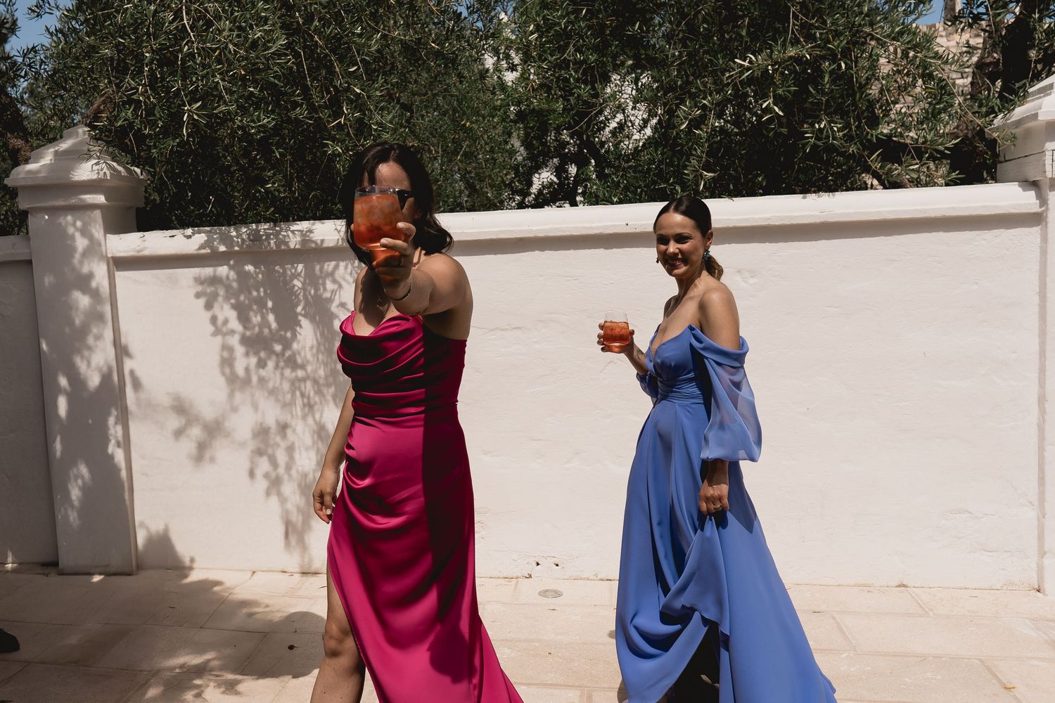 Two women in colorful formal dresses enjoying drinks at outdoor event.