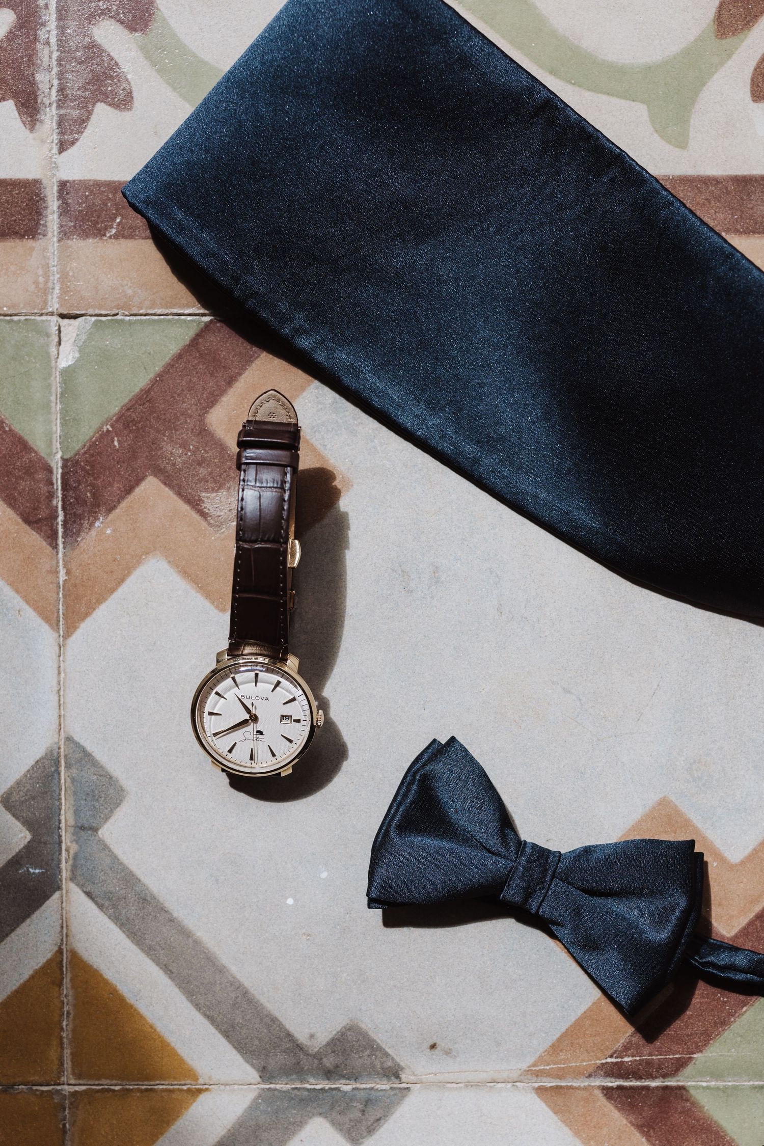 Wedding accessories including wristwatch and navy blue bow tie arranged on patterned tile floor.