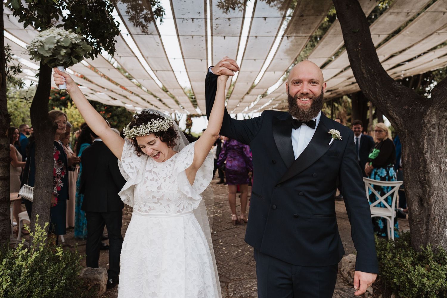 Jubilant newlyweds raise hands in celebration while exiting ceremony under draped canopy.