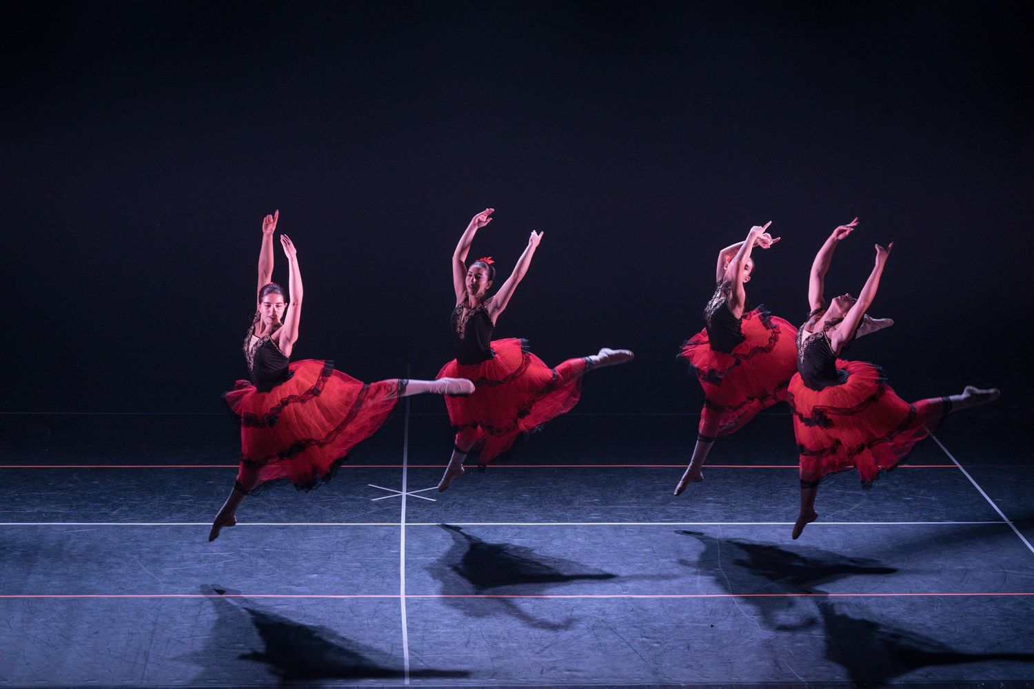 Ballet dancers in flowing red dresses leap gracefully across stage in synchronized motion.