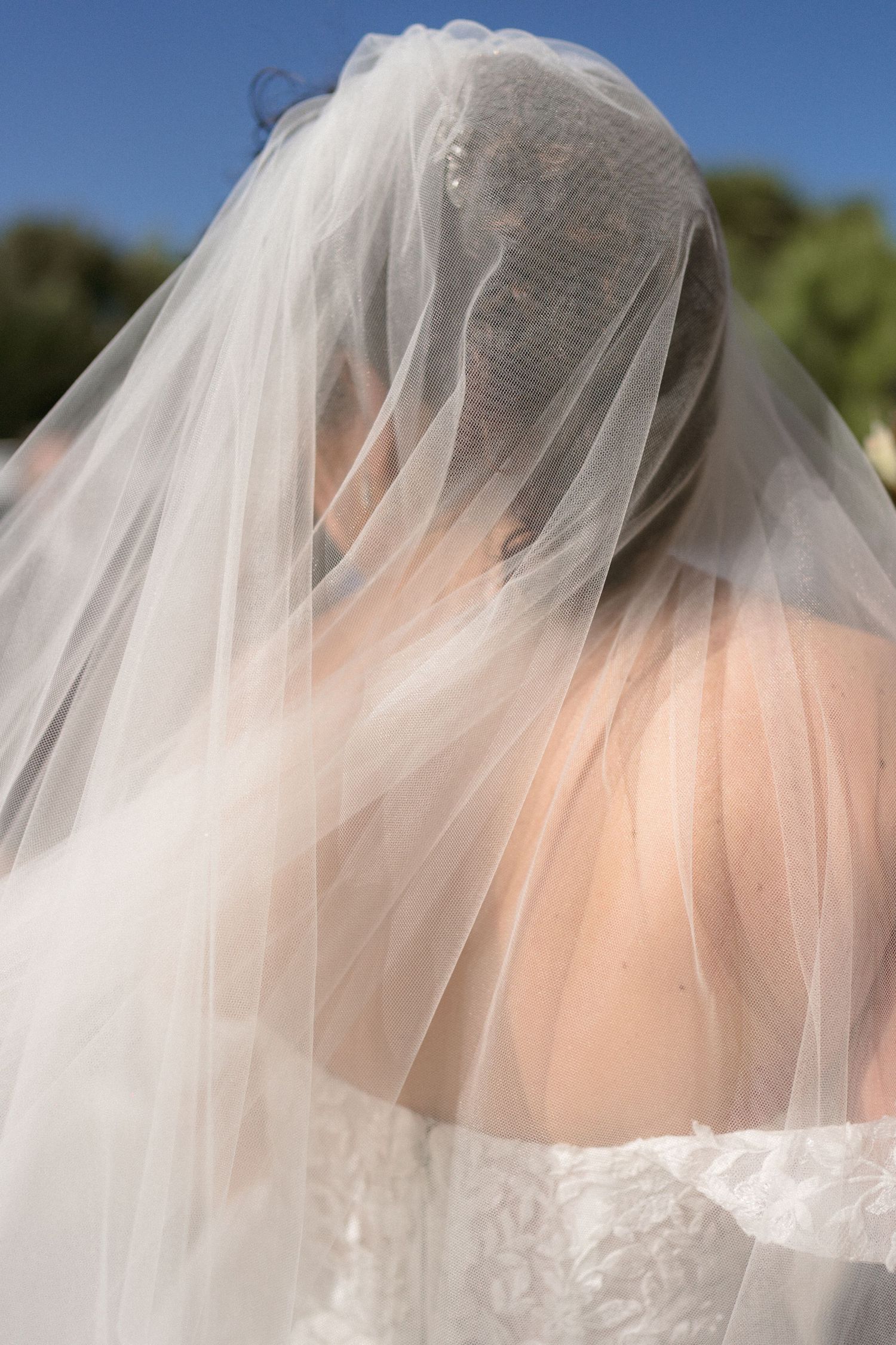 Bride wearing a flowing white wedding veil draped over her head outdoors on a sunny day.