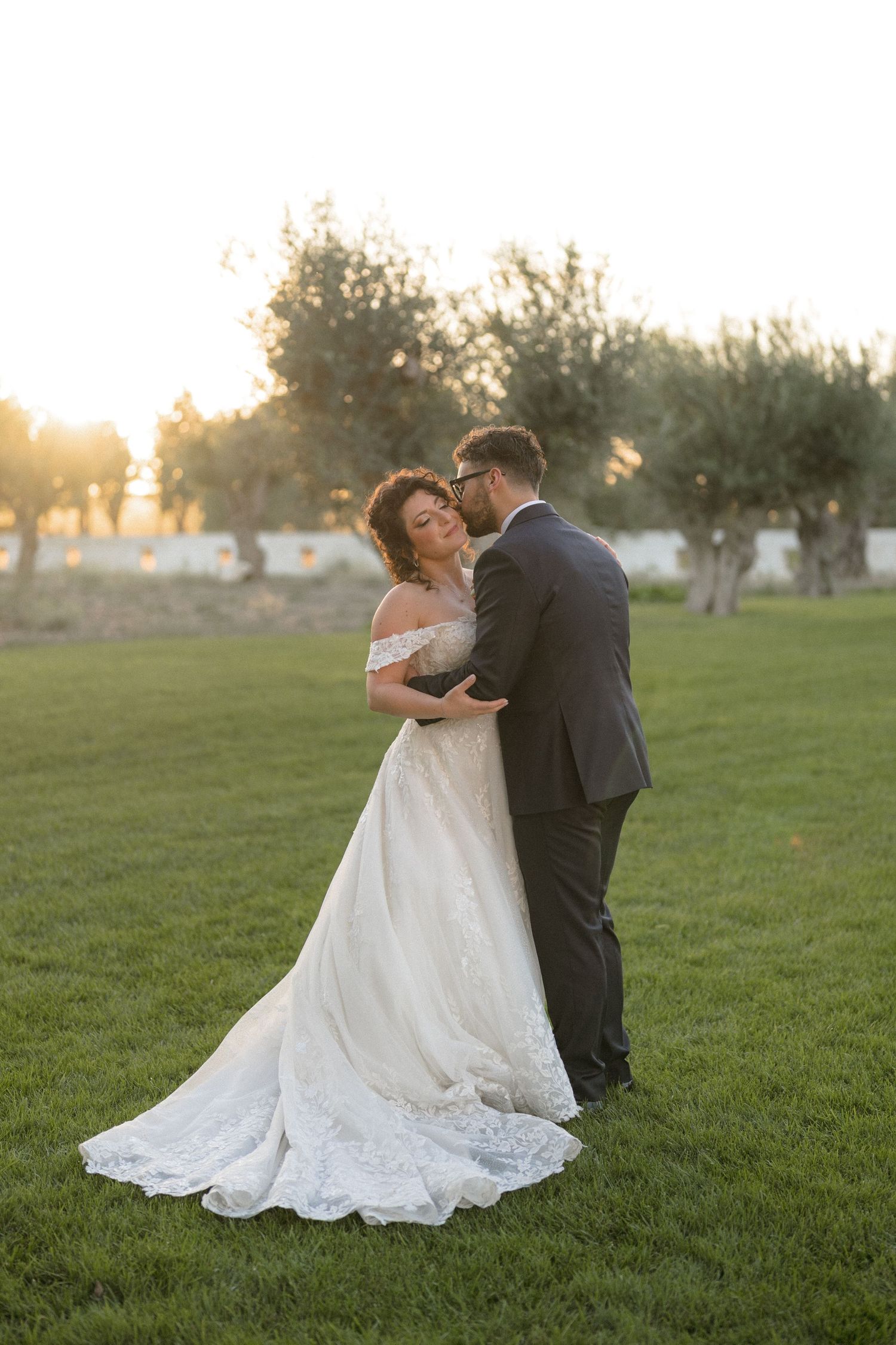 Bride and groom embracing at golden hour sunset in a lush green garden with olive trees in the background.