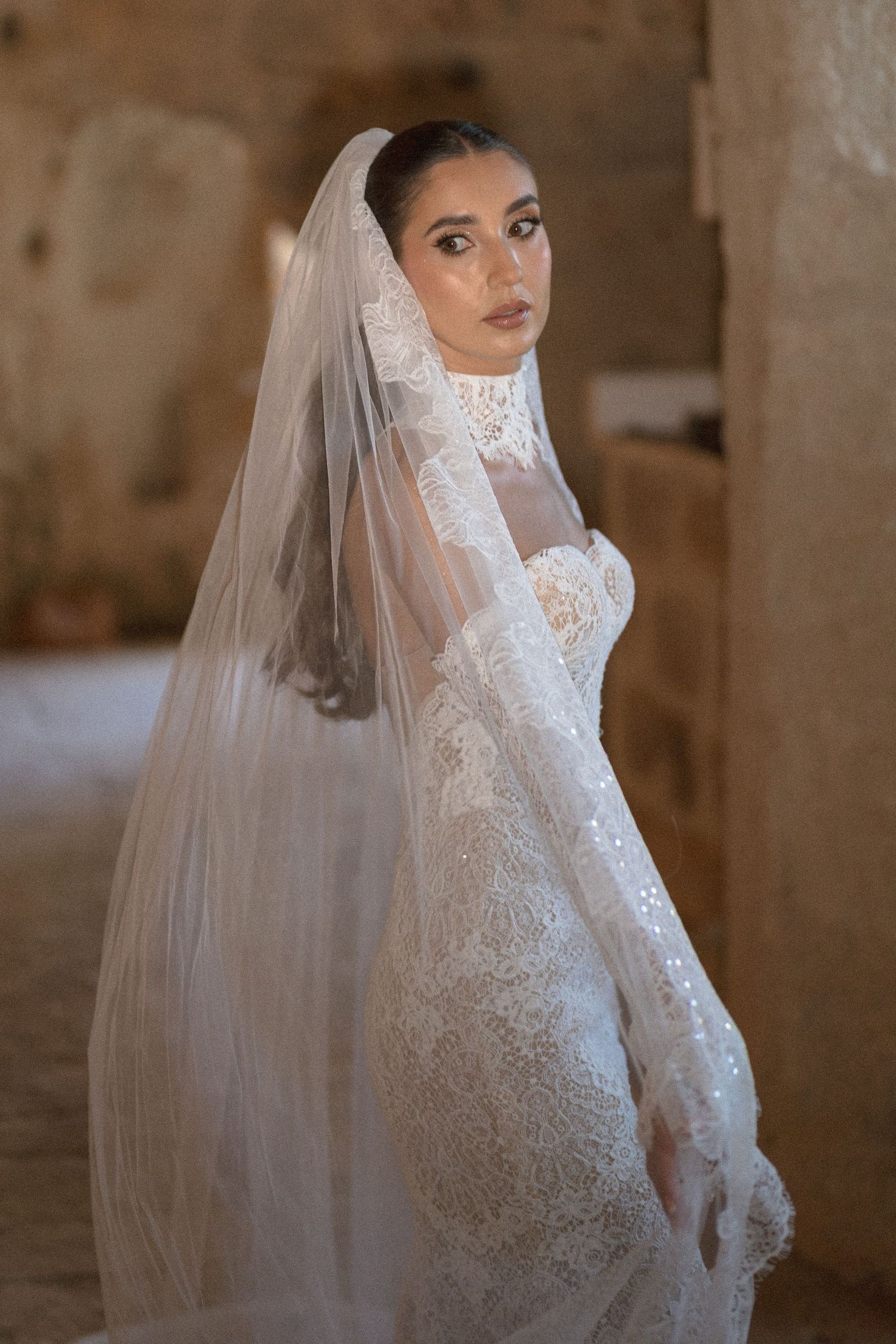 Bride in a fitted lace wedding gown and long sheer veil looks over her shoulder inside a warm, rustic stone interior.
