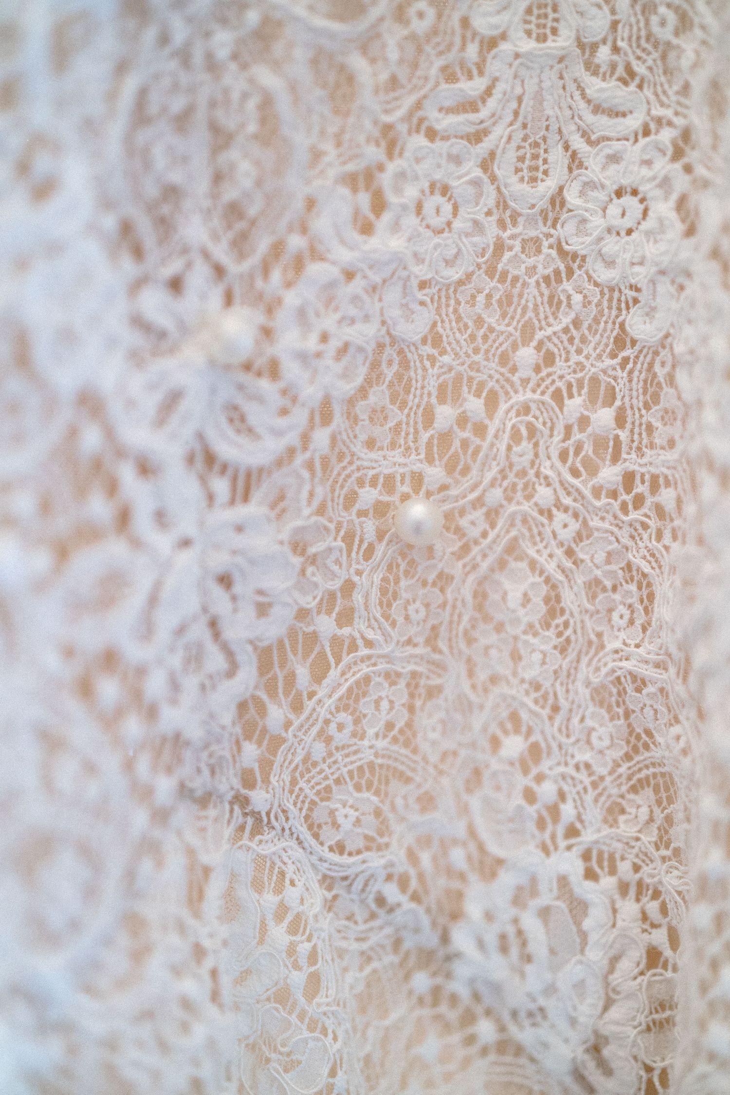 Close-up of delicate white floral lace fabric with pearl accents over a soft blush underlay.