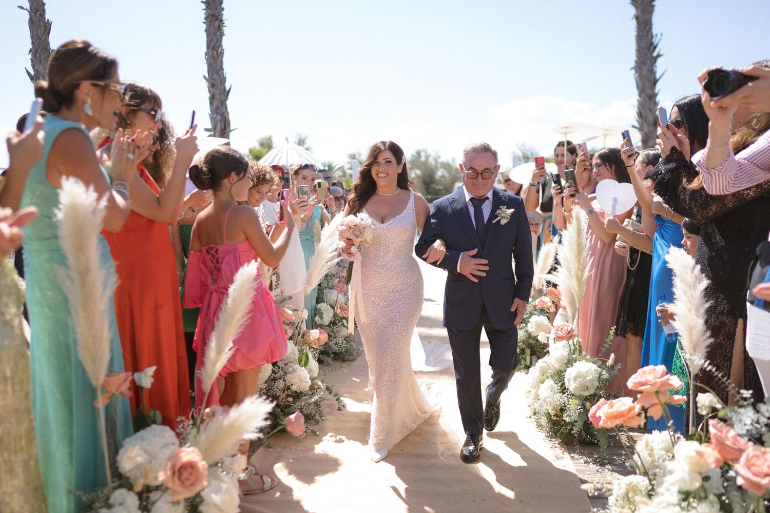 Outdoor beach wedding ceremony with guests in pastel attire and palm trees.