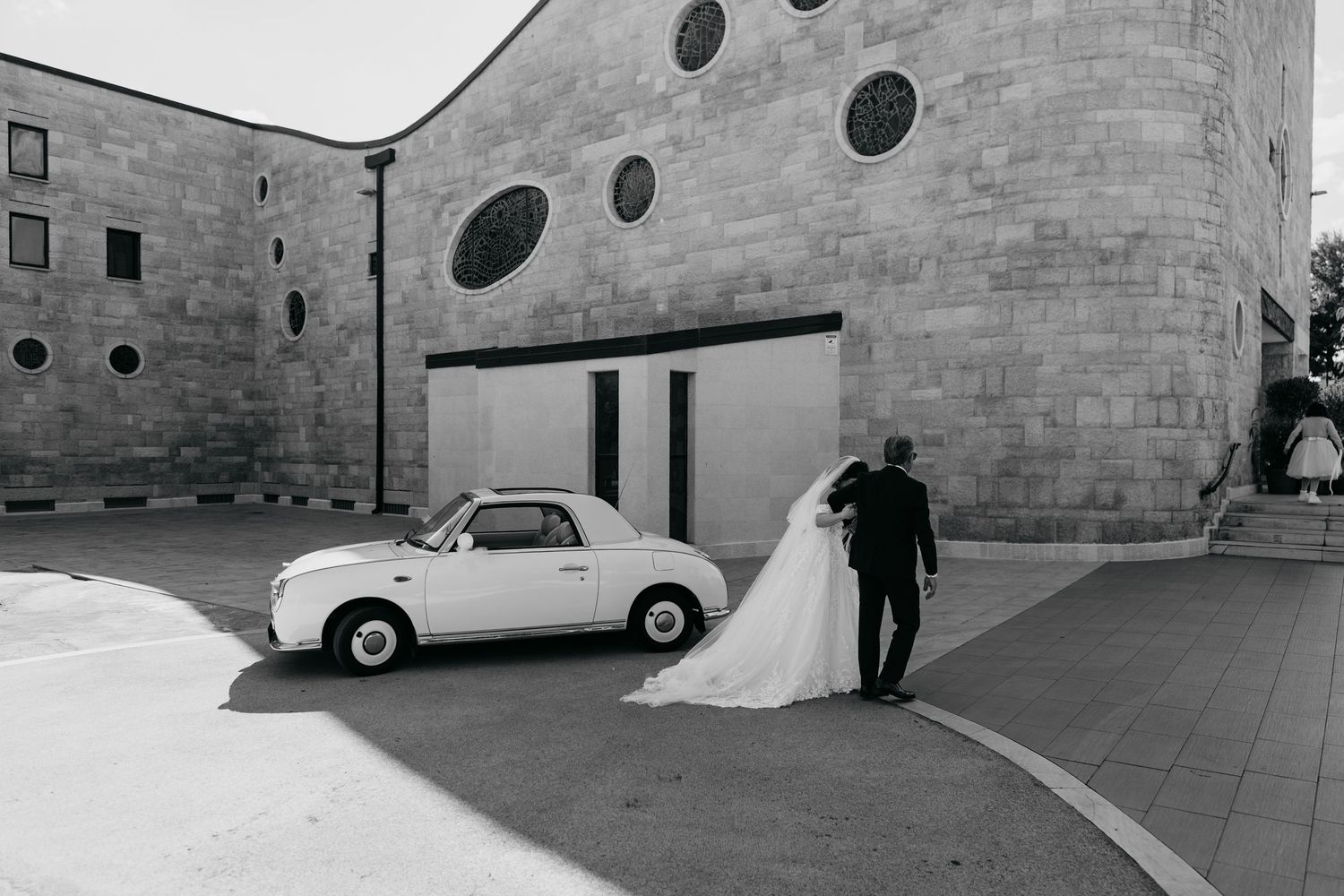 Bride and groom stand beside a vintage white car outside a stone church in a black and white wedding photo.