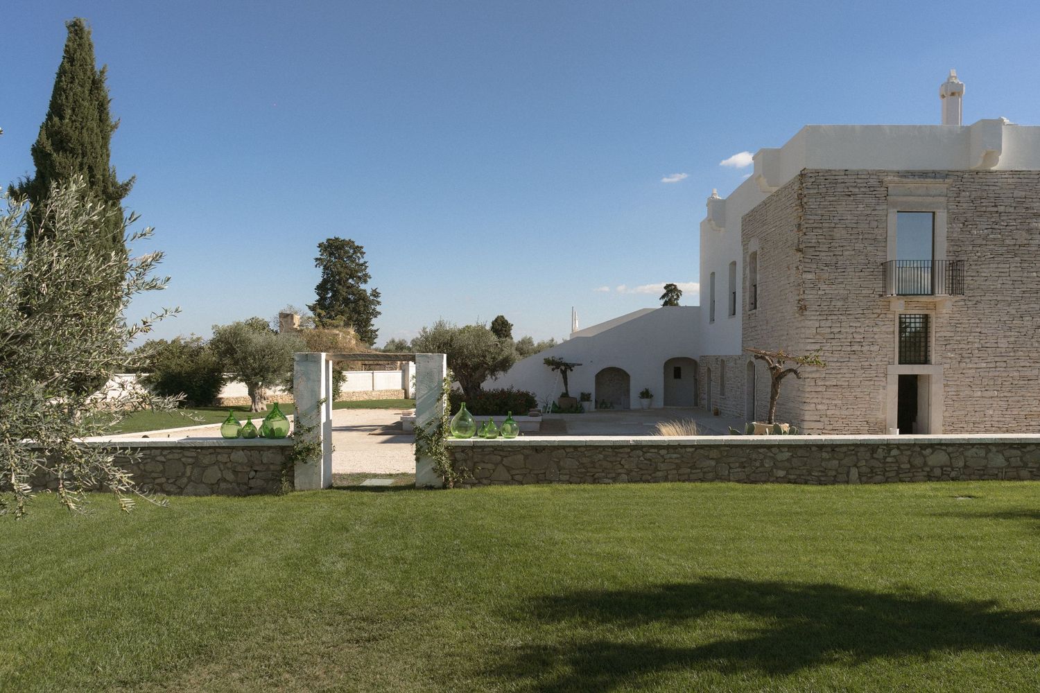 Elegant white stone wedding venue with lush green lawn, cypress trees, and rustic architecture under blue sky.