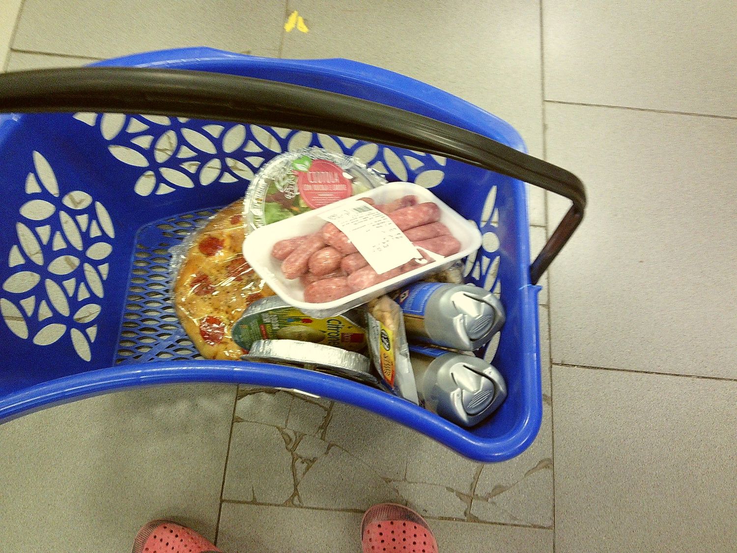 Shopping basket containing groceries and household items on gray floor tiles.