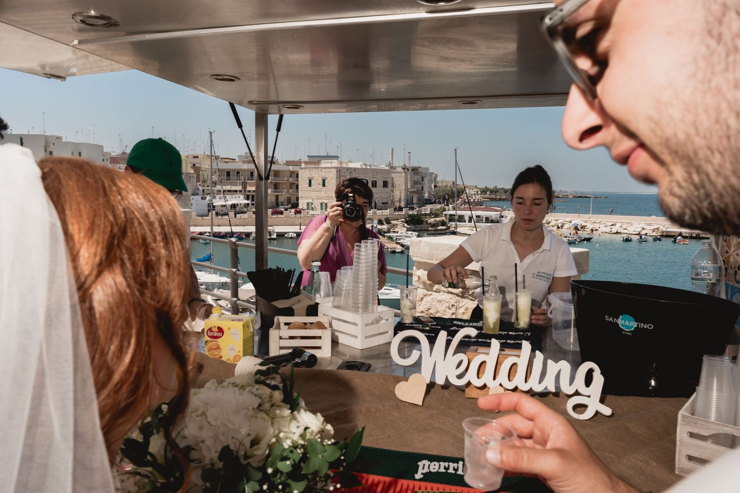 People celebrate at an outdoor wedding reception venue overlooking a Mediterranean harbor in summer.