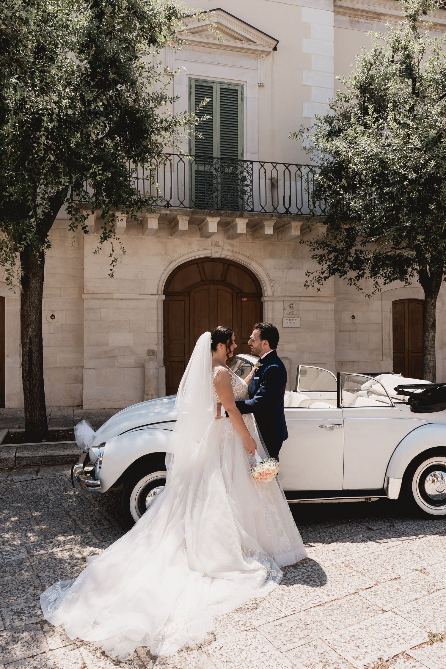 A romantic wedding moment next to a vintage white car on a Mediterranean street.