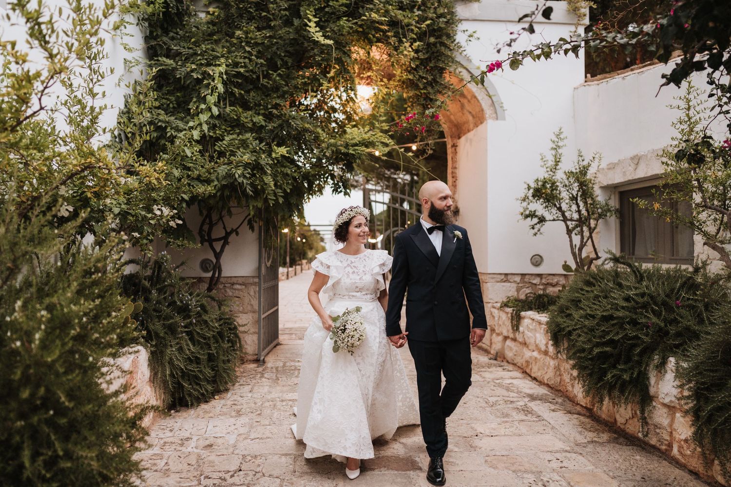 A wedding couple walking along a sunlit garden pathway beside white Mediterranean architecture.