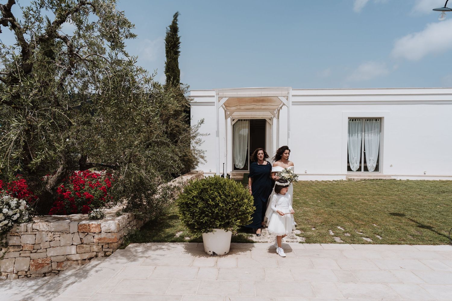 Wedding couple embraces outside Mediterranean-style white building with flowering bushes.