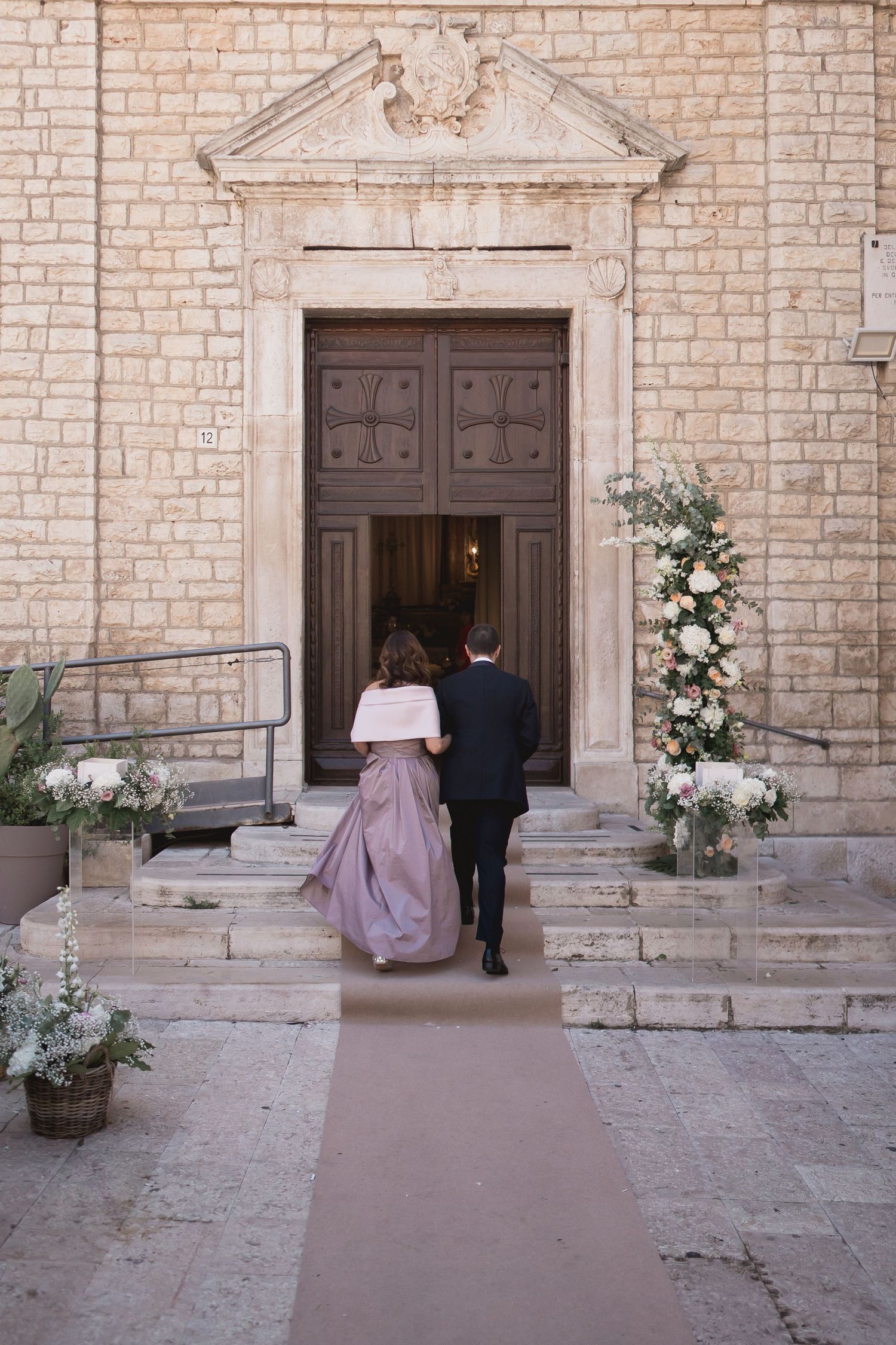 Couple entering stone church doorway decorated with white floral arrangements for wedding ceremony.