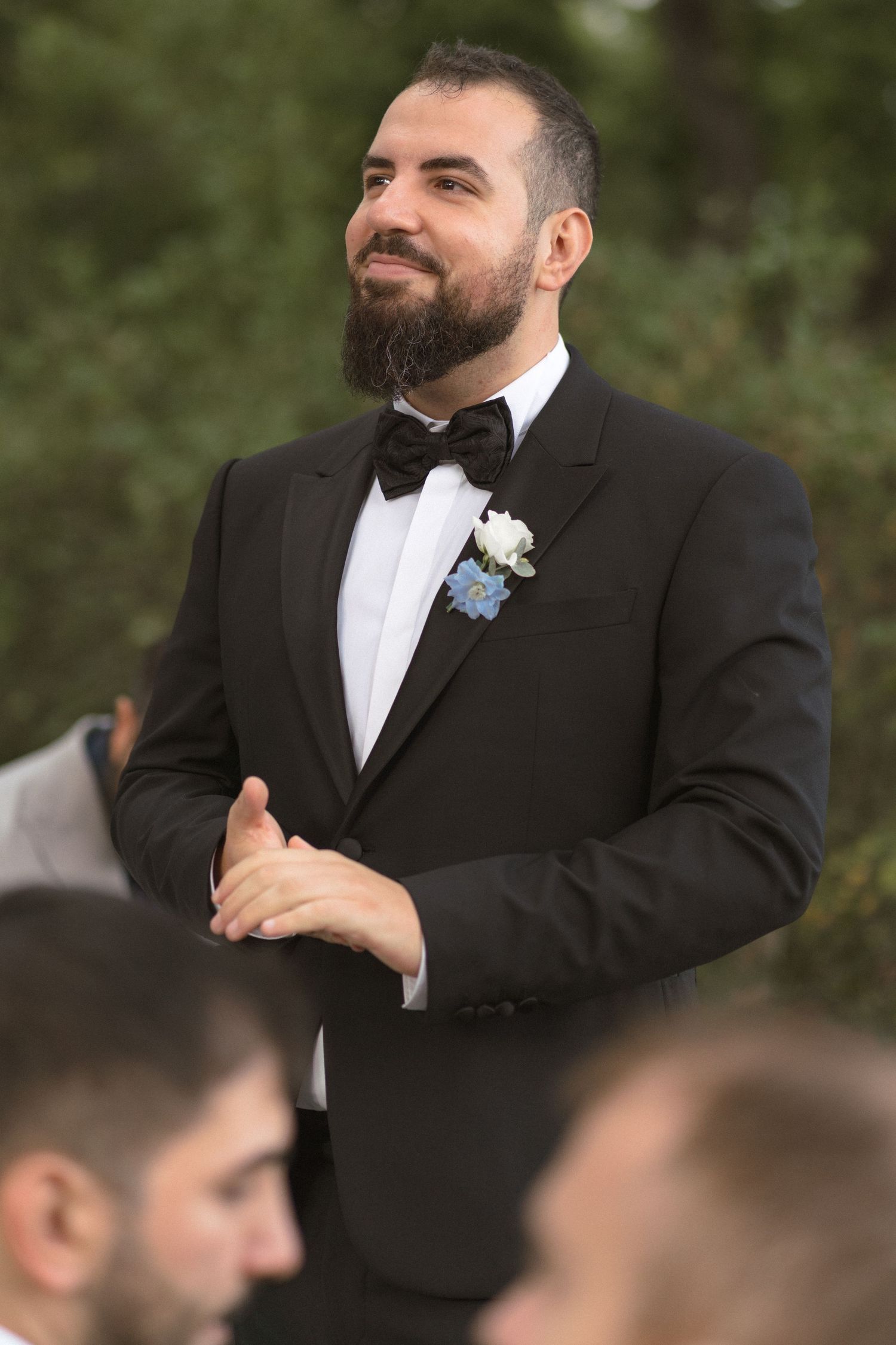 A groomsman in a black tuxedo and white boutonniere smiles during an outdoor wedding ceremony.