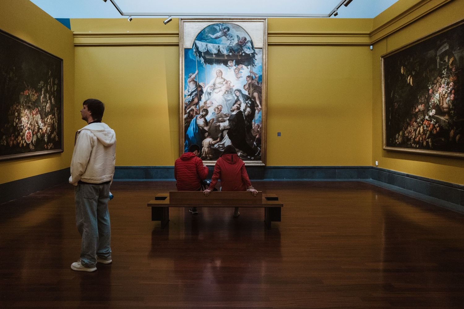 Museum visitors admire large classical paintings in an elegant gallery with yellow walls and polished wooden floors.