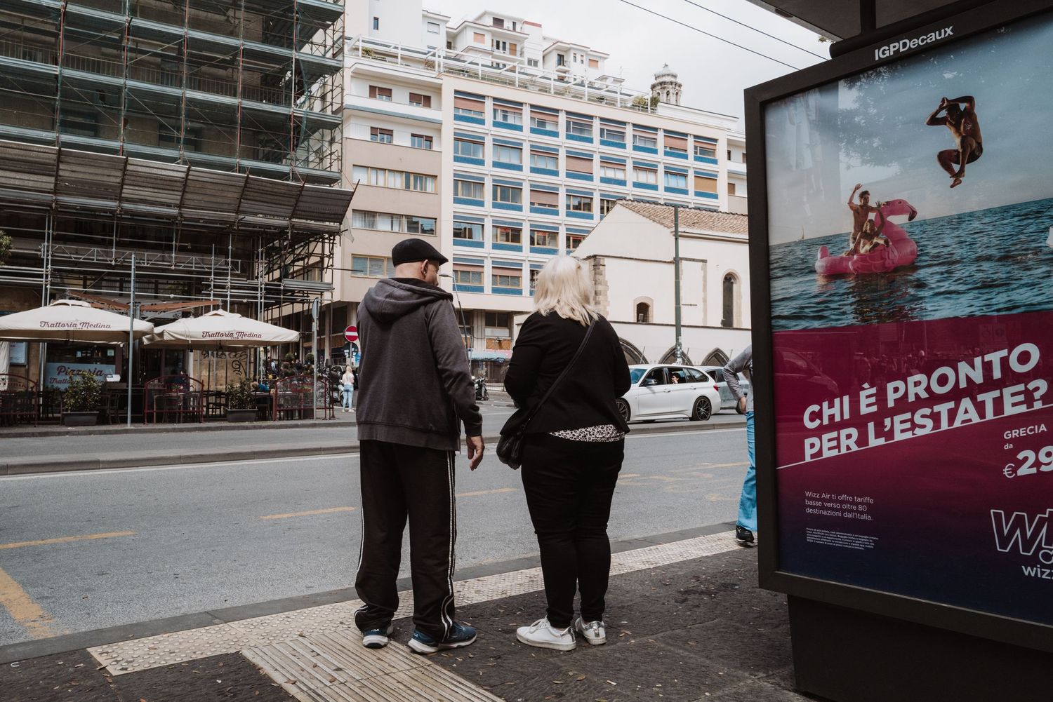 Two people wait at a bus stop in an urban setting with scaffolding and a digital advertisement display nearby.
