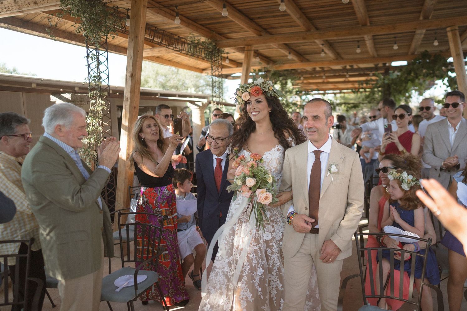 Outdoor wedding ceremony under a wooden pergola decorated with greenery.