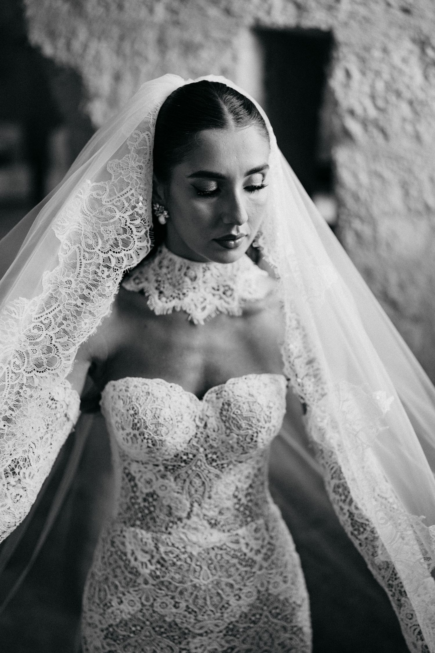 Black and white portrait of a bride looking down, wearing a strapless lace gown and a lace-edged veil.