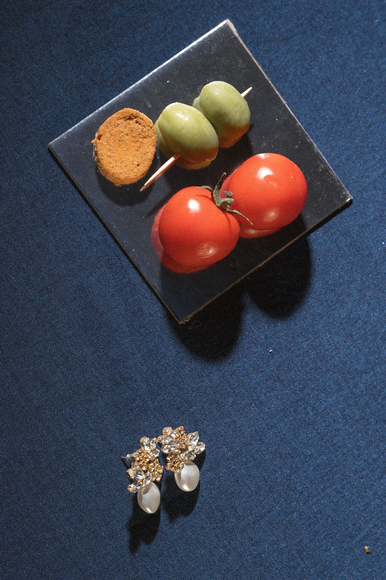 Two ripe tomatoes, olive skewers and a bread slice sit on a dark slate plate above floral pearl earrings on blue fabric.