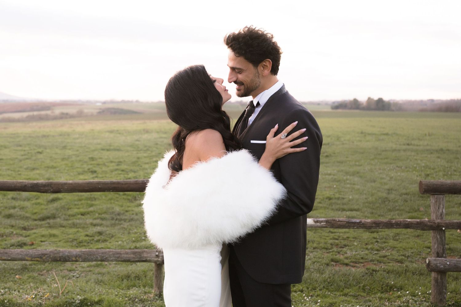 A bride and groom share a romantic moment in a white fur stole and dark suit at an outdoor farm wedding.