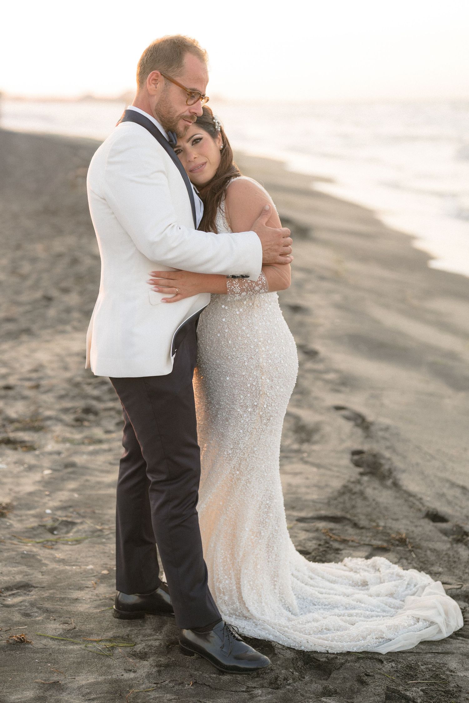 A romantic beach wedding photo of a couple embracing on the sand at sunset, with the bride in a sparkly white gown.