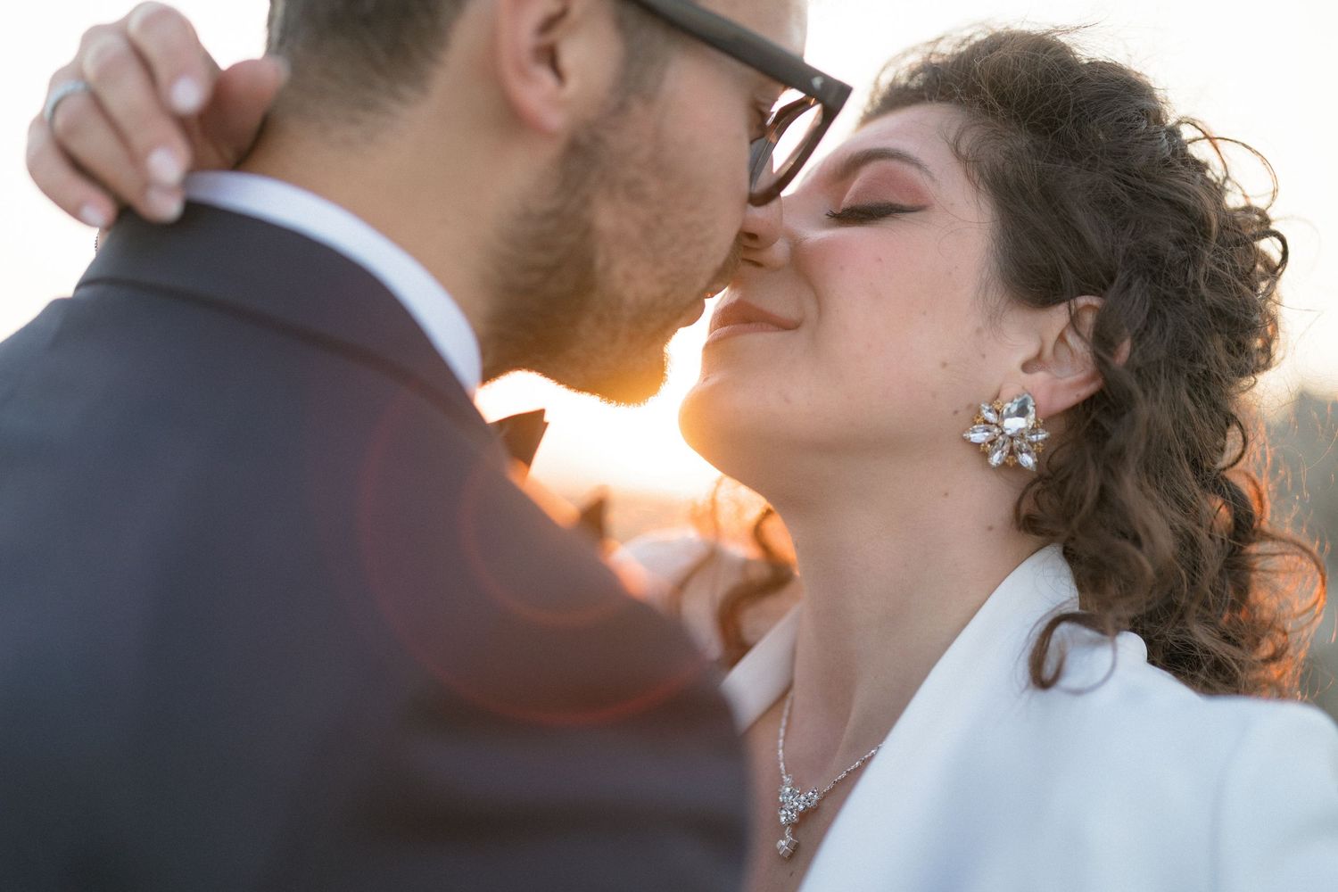 Couple sharing a romantic kiss at sunset, bride wearing floral earrings and white jacket, groom in gray suit.