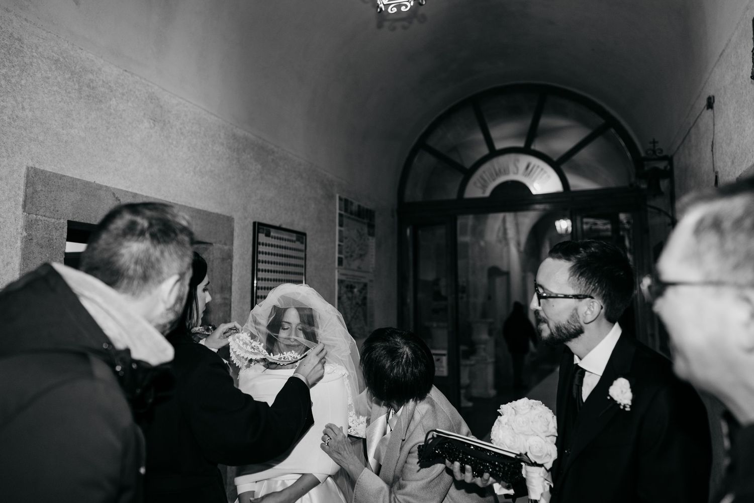 Bride surrounded by guests in black and white photo inside arched corridor after wedding ceremony.