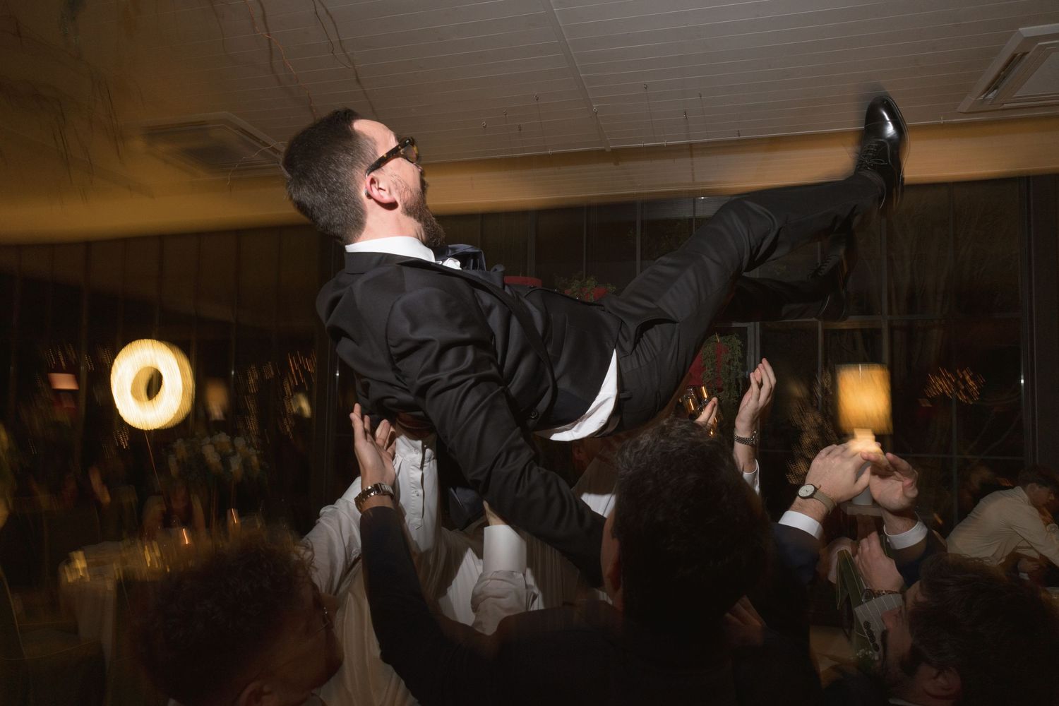 Groom in black tuxedo being lifted by guests during lively wedding reception celebration.