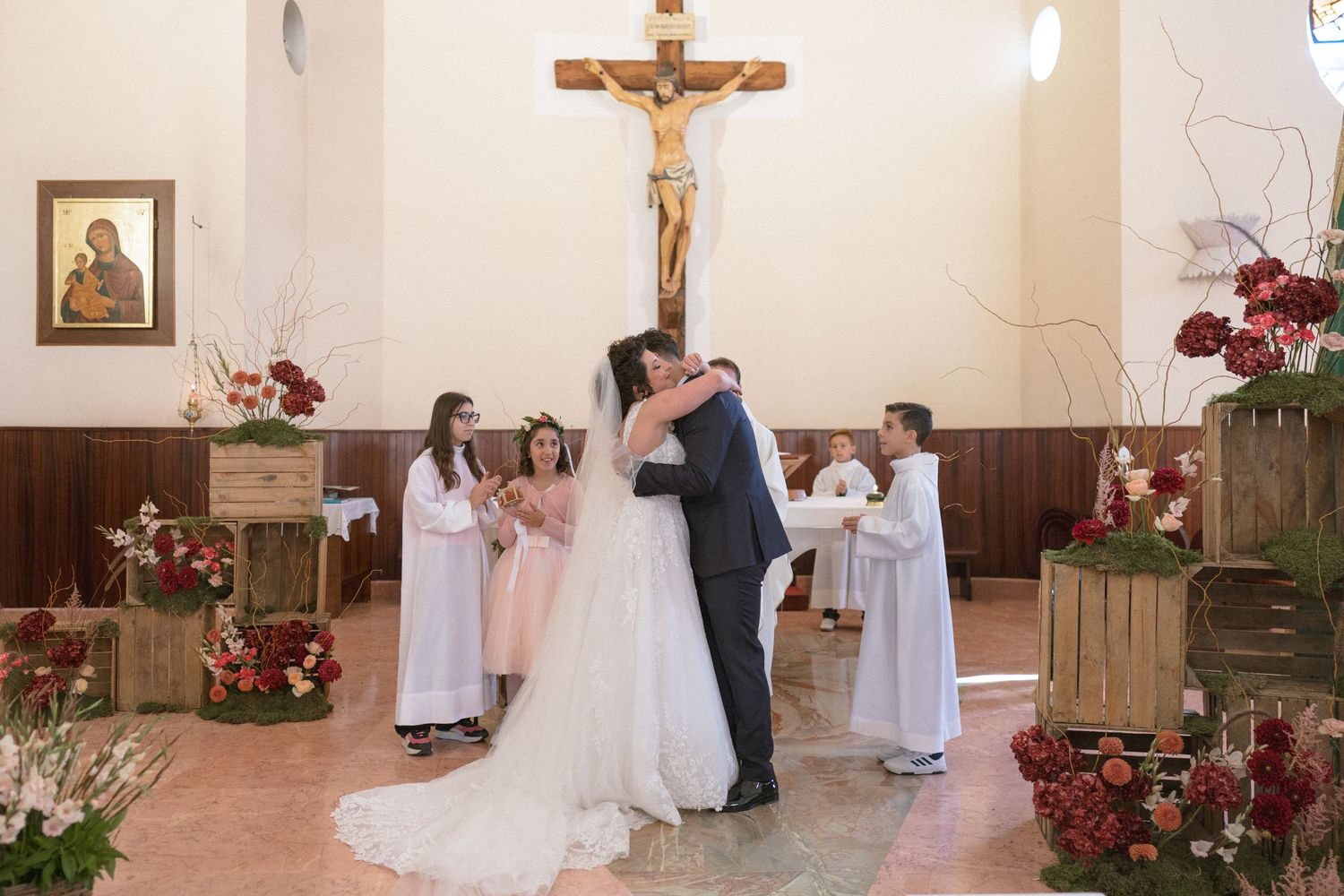Bride and groom share first kiss at altar decorated with red flowers and wooden crates in church.