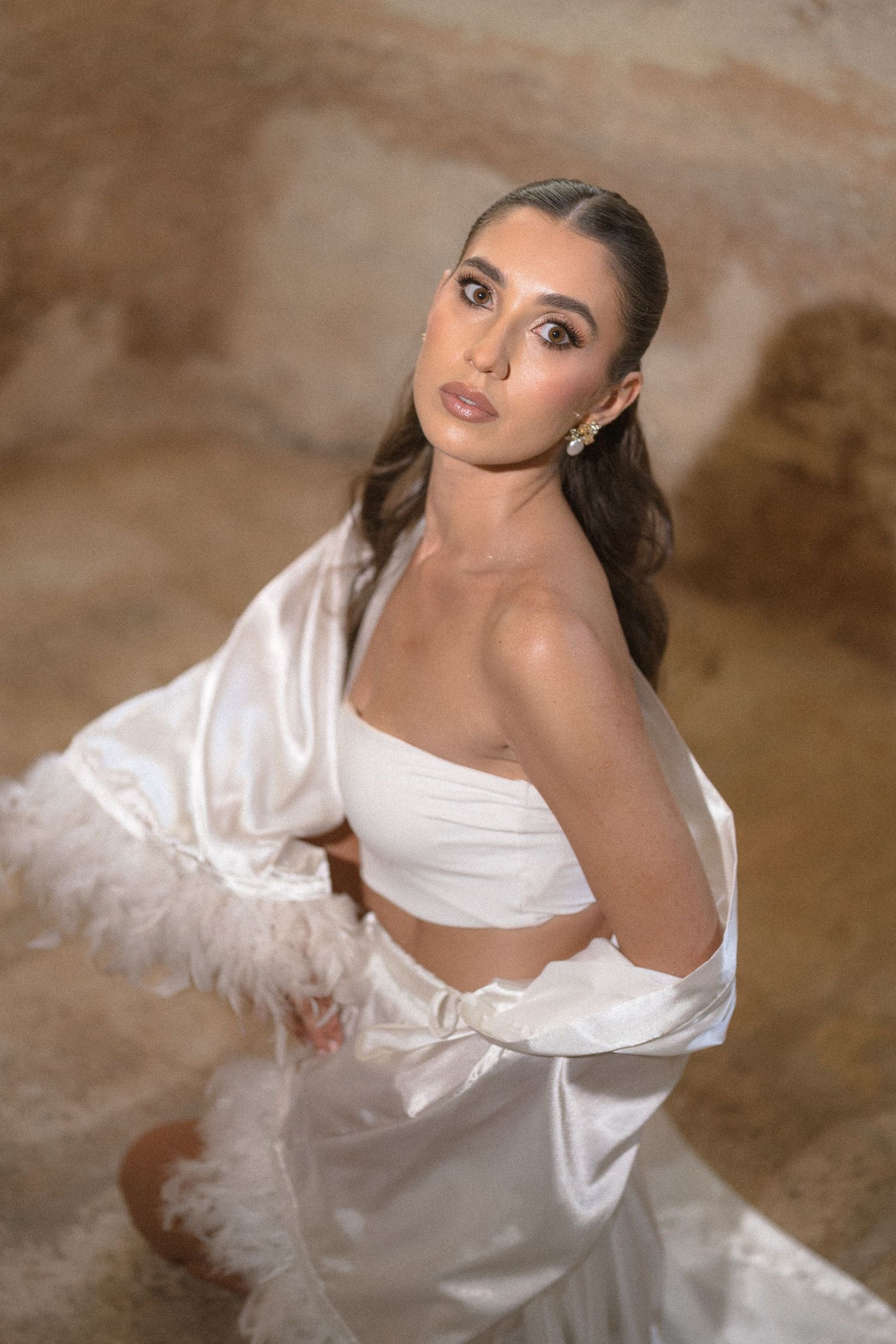 Bride with sleek hair and drop earrings looks up at the camera in a strapless white satin outfit with feather trim.