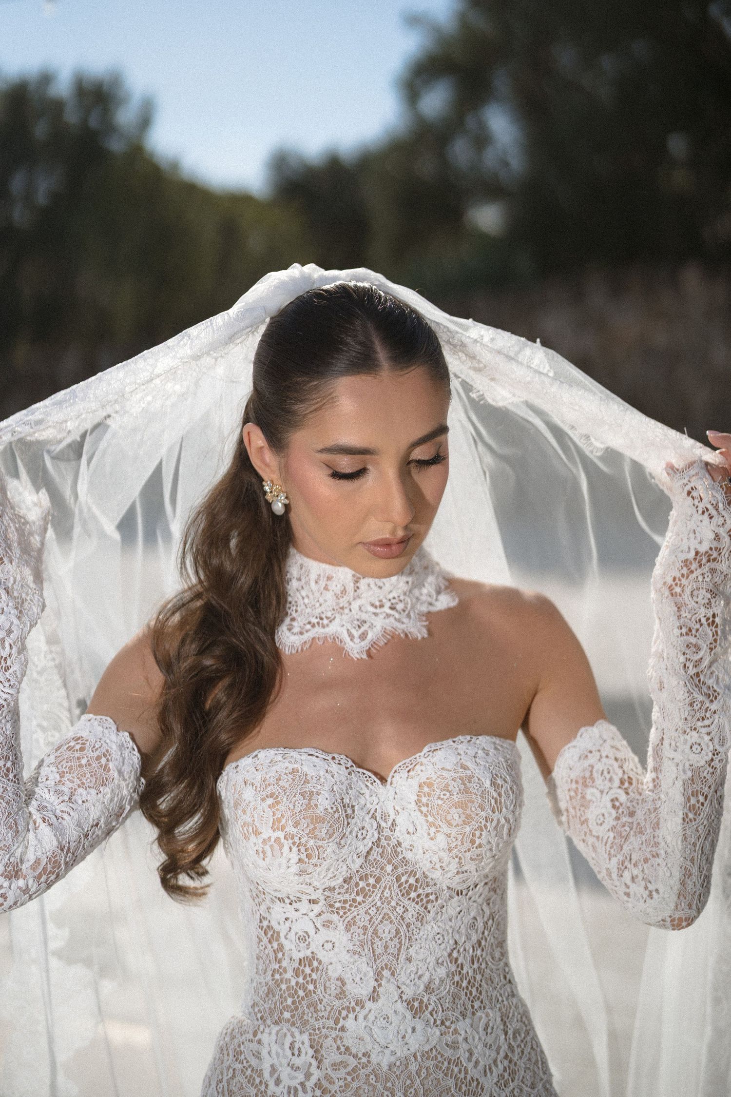 Bride with long wavy hair lifts her sheer lace veil, wearing a lace gown with sheer sleeves and a choker-style collar outdoors.