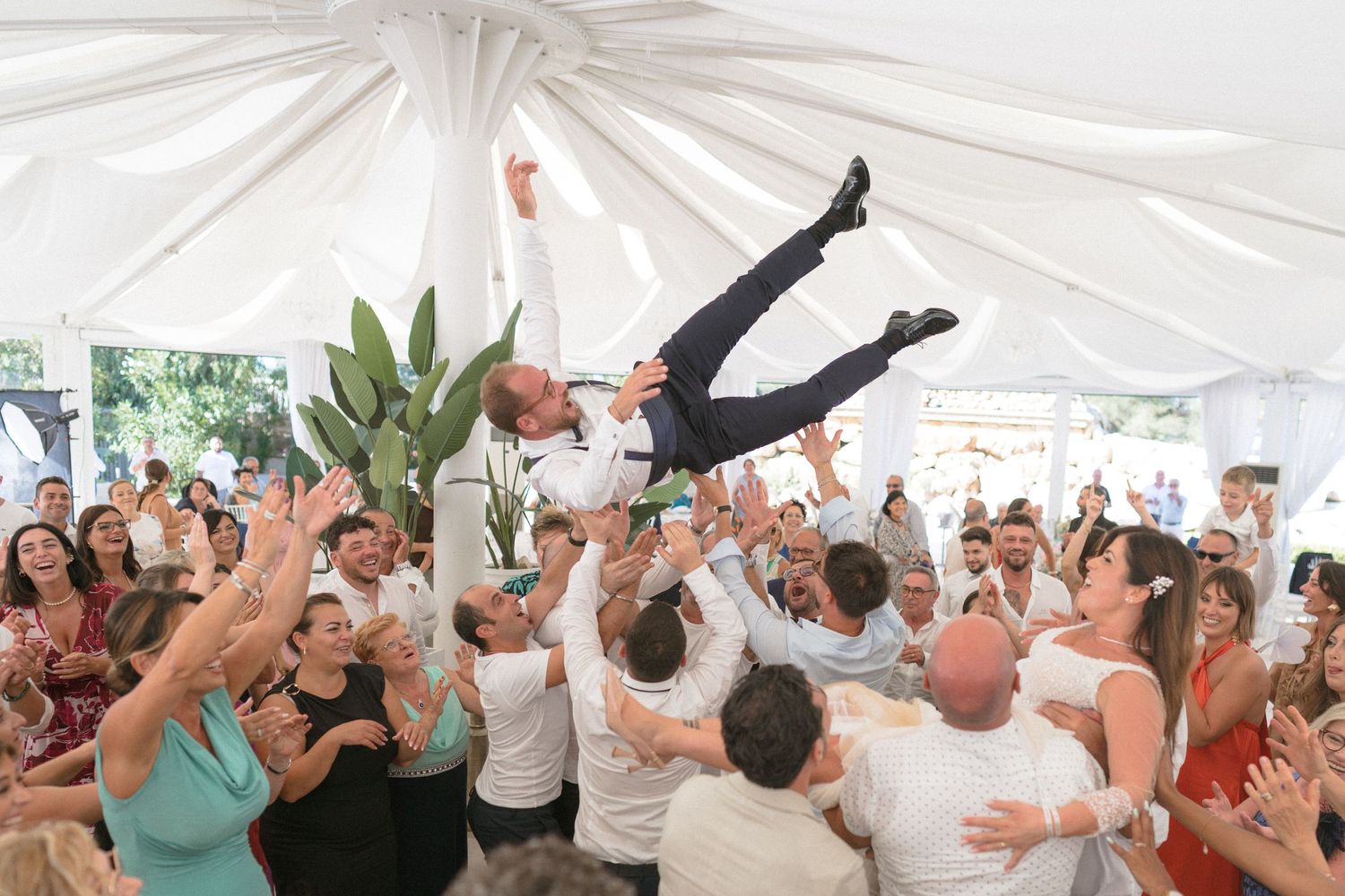 Wedding guests toss and lift someone in celebration under a white tent during a lively reception party.