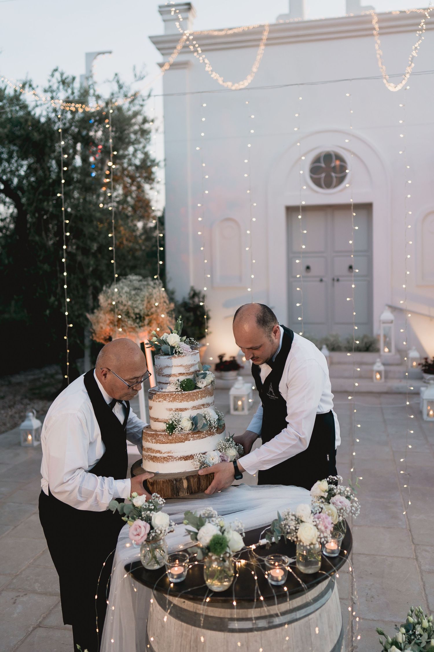 Two catering staff members arranging a tiered wedding cake outdoors with string lights and white building backdrop.