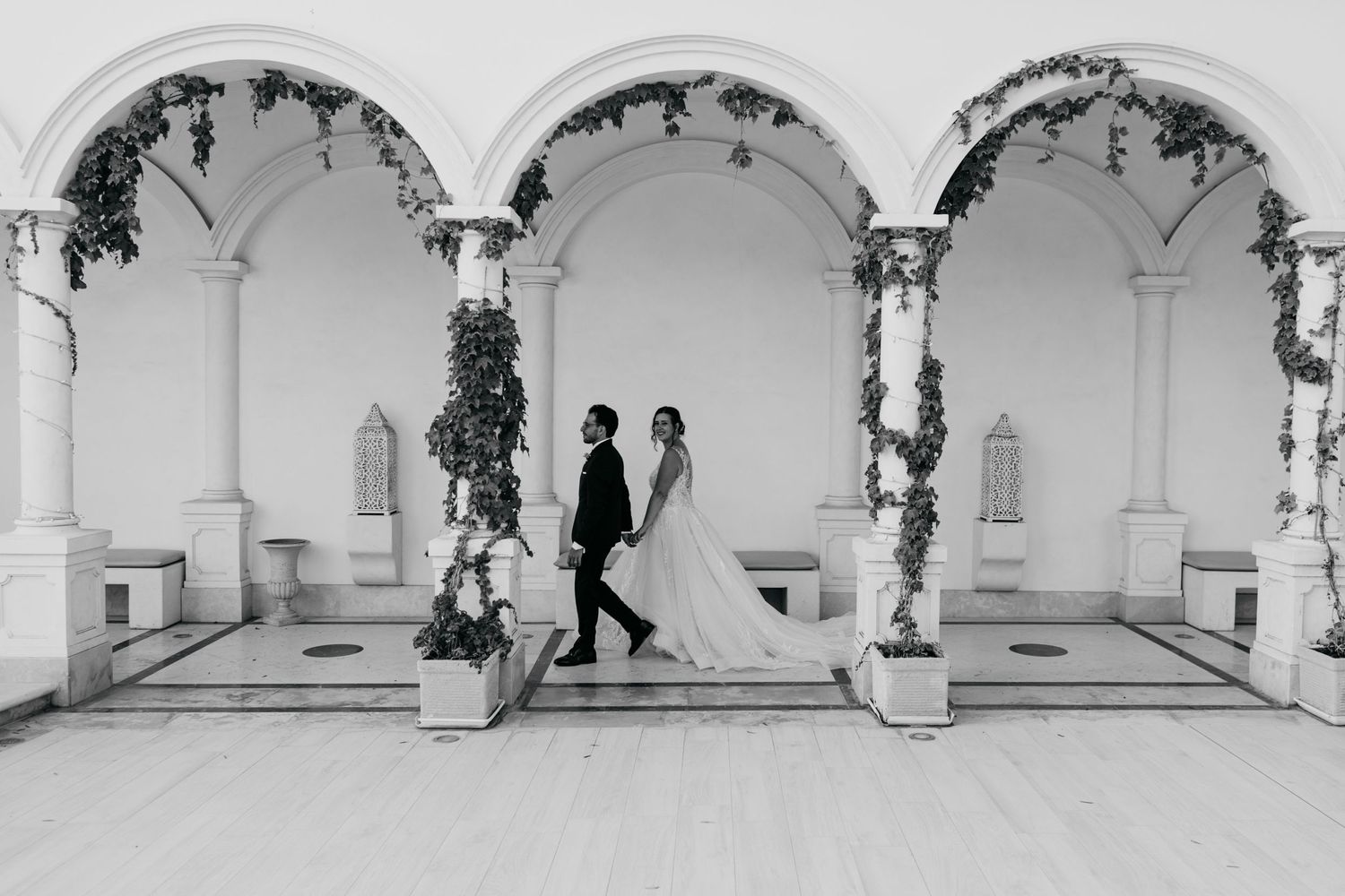 Black and white photo of couple walking through elegant archway with vine covered columns.