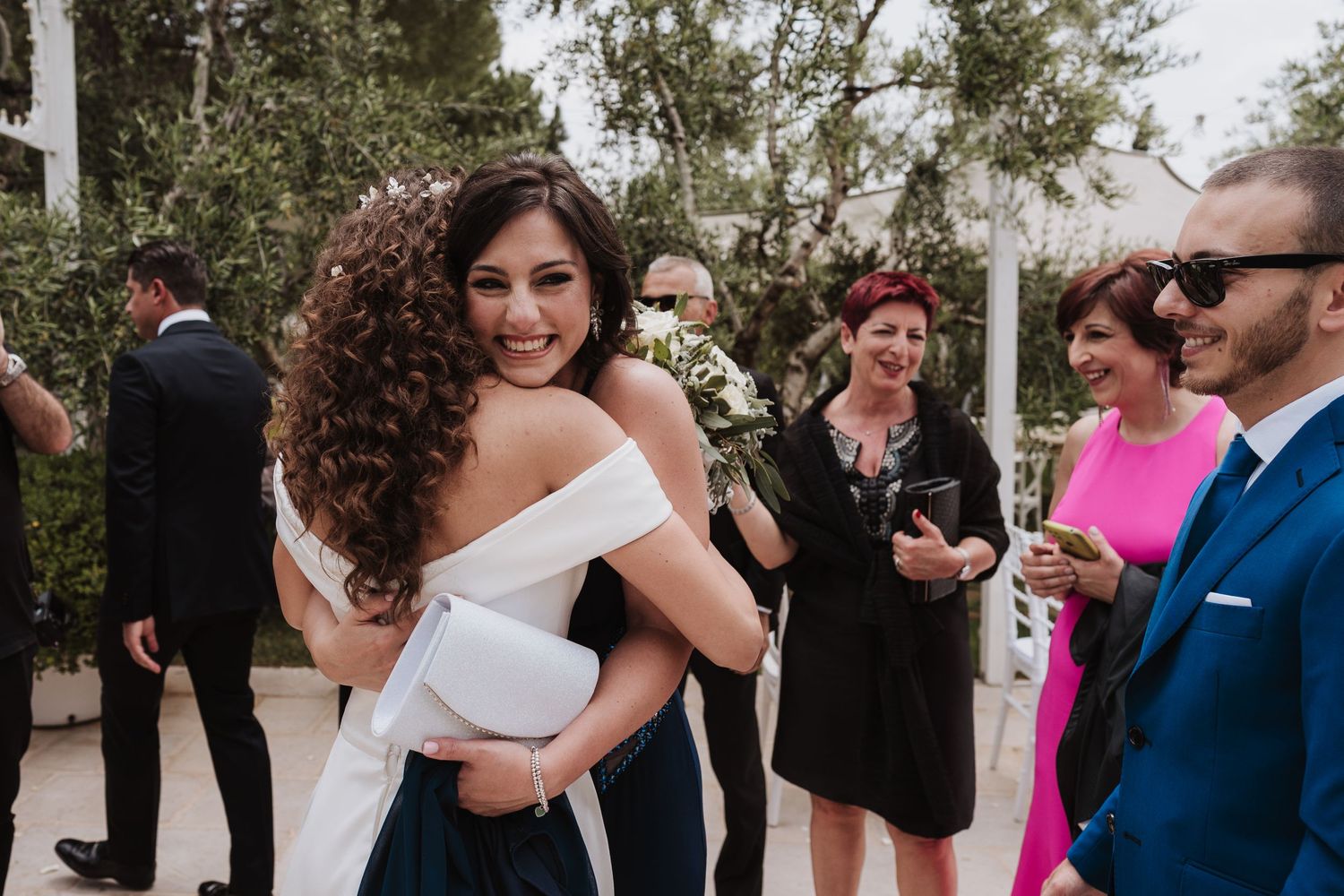 Wedding guests celebrate and embrace during an outdoor reception with olive trees in the background.