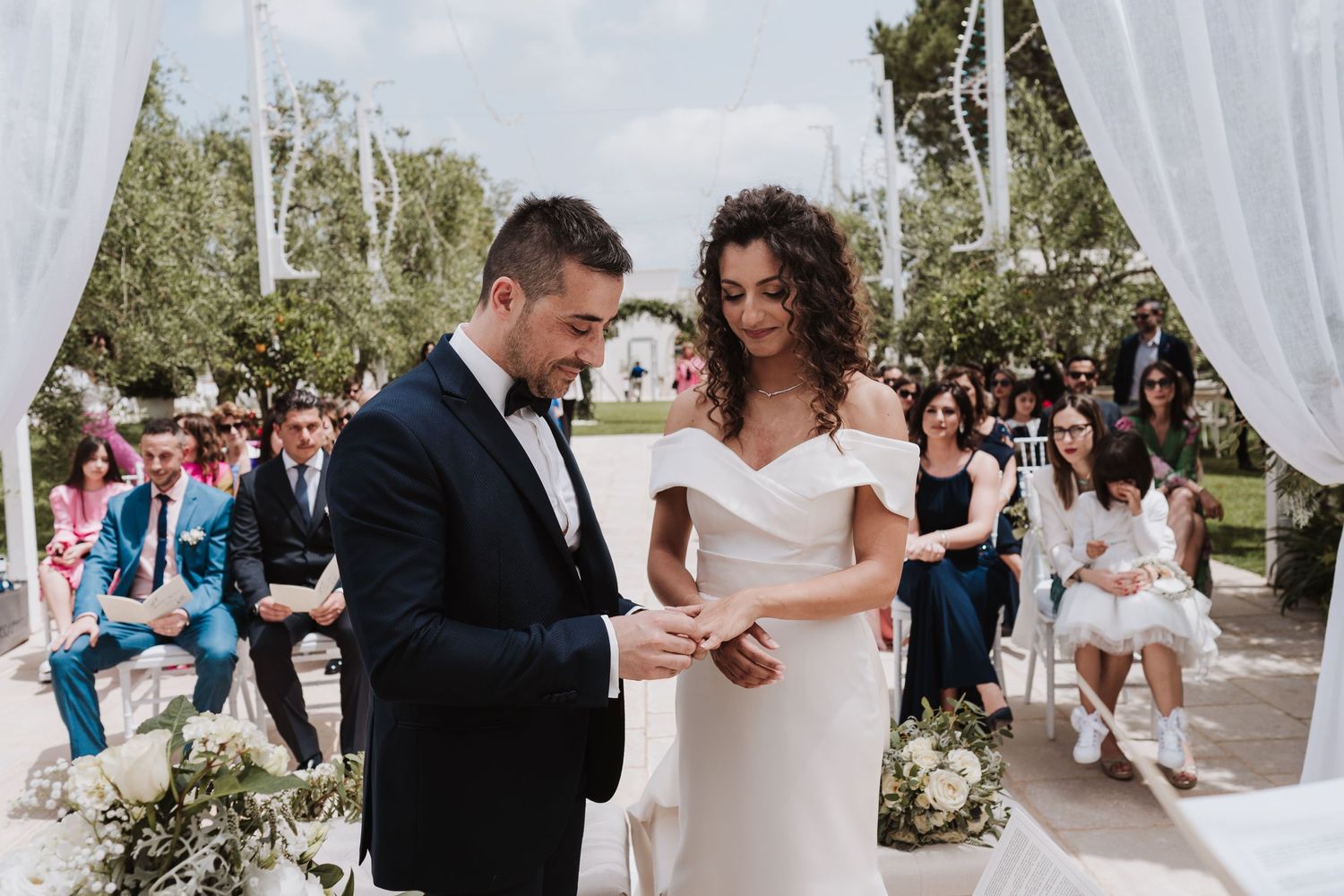 Wedding ceremony exchange of rings under white drapes with guests seated in background.