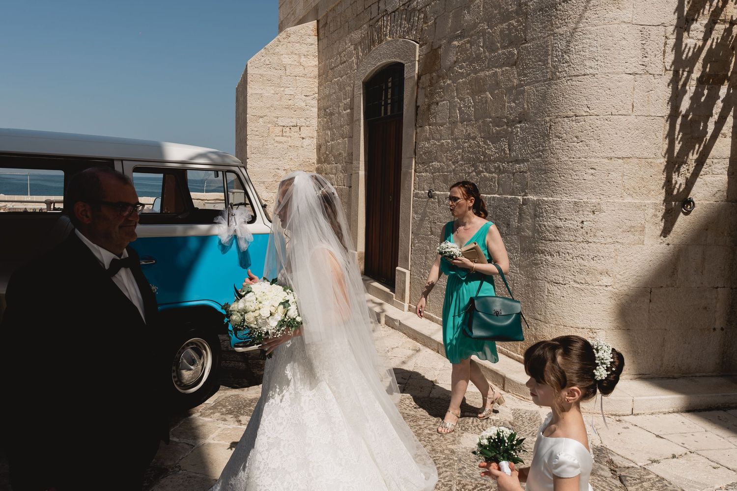 Wedding party members in turquoise dresses assist with a flowing white veil outdoors.