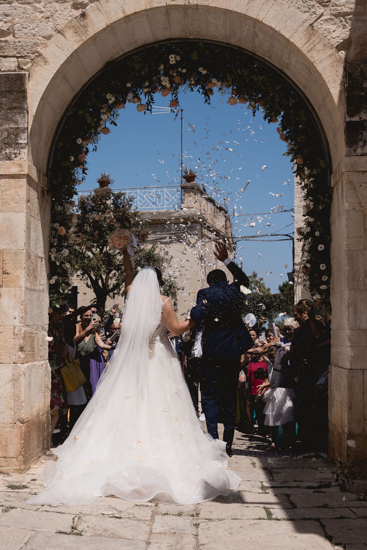 A wedding ceremony exit through a stone archway as guests shower confetti.