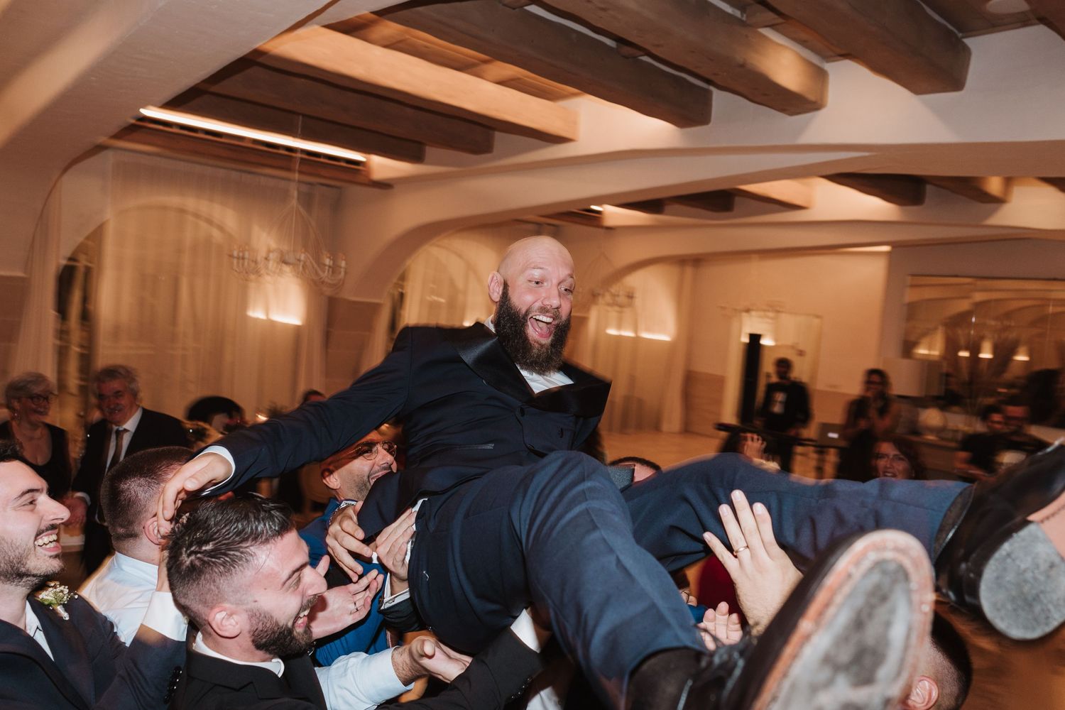 Wedding guests lift a person in formal attire during an energetic reception celebration.
