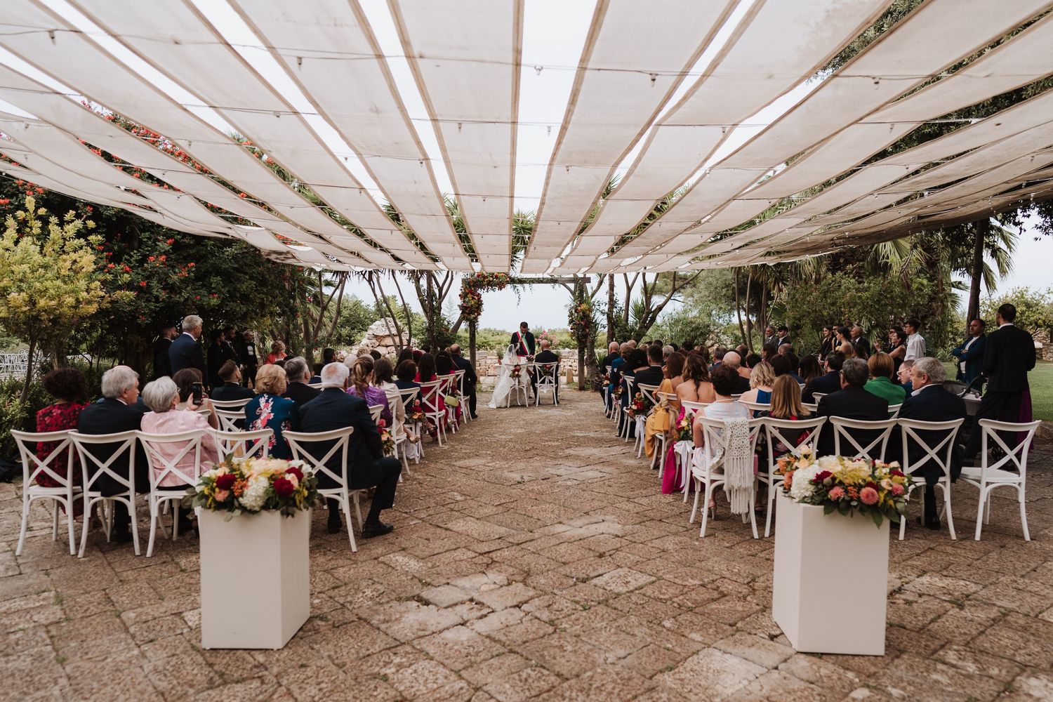 Outdoor wedding venue with white canopy, cross-back chairs, and floral arrangements lining the aisle.