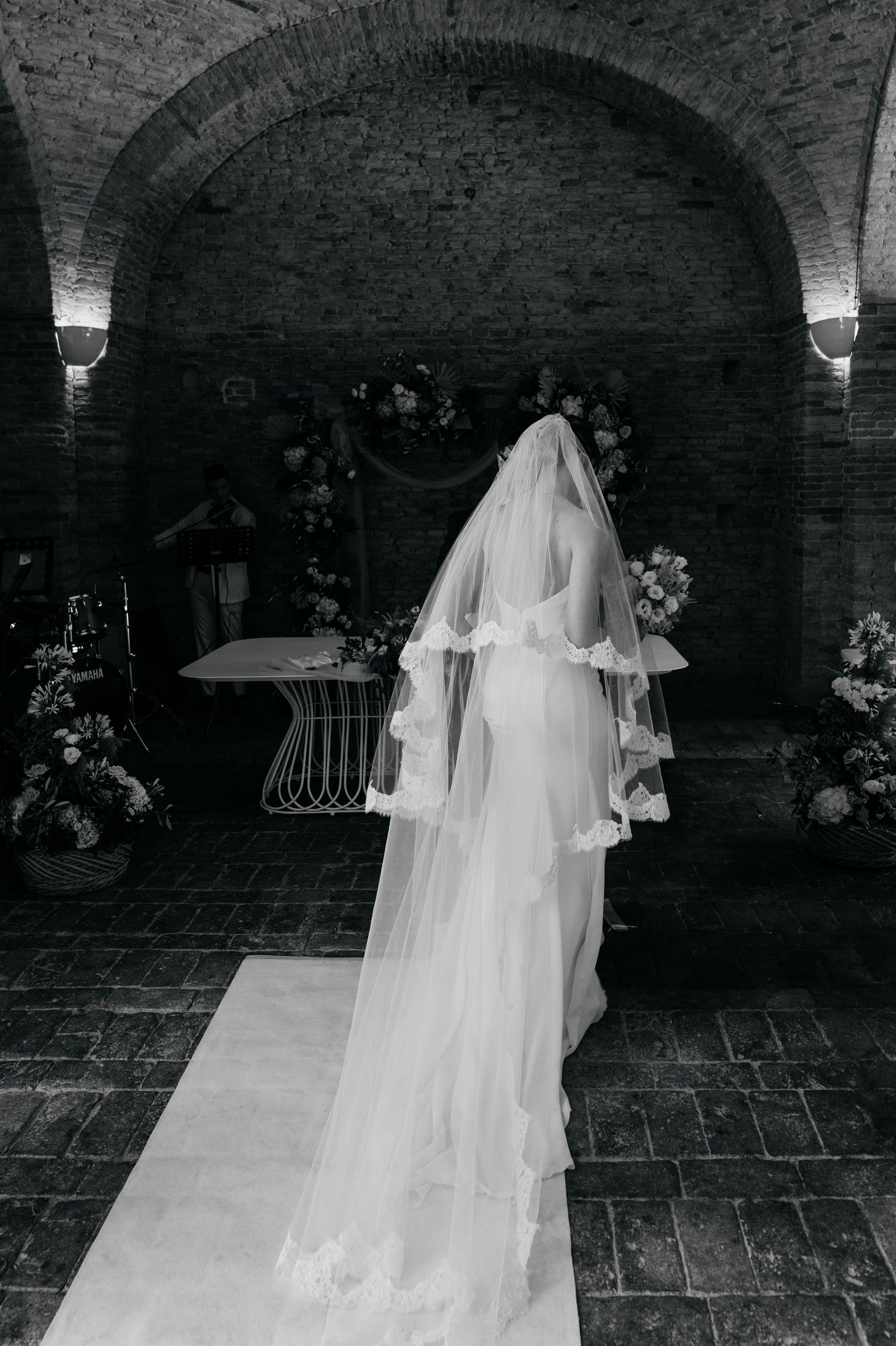 Bride in flowing white dress and veil walking through elegant brick archways.