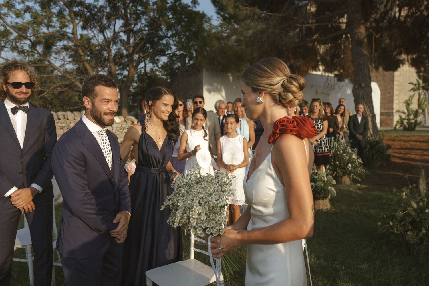 A group gatheres outdoors for an elegant wedding ceremony under tall trees on a sunny day.