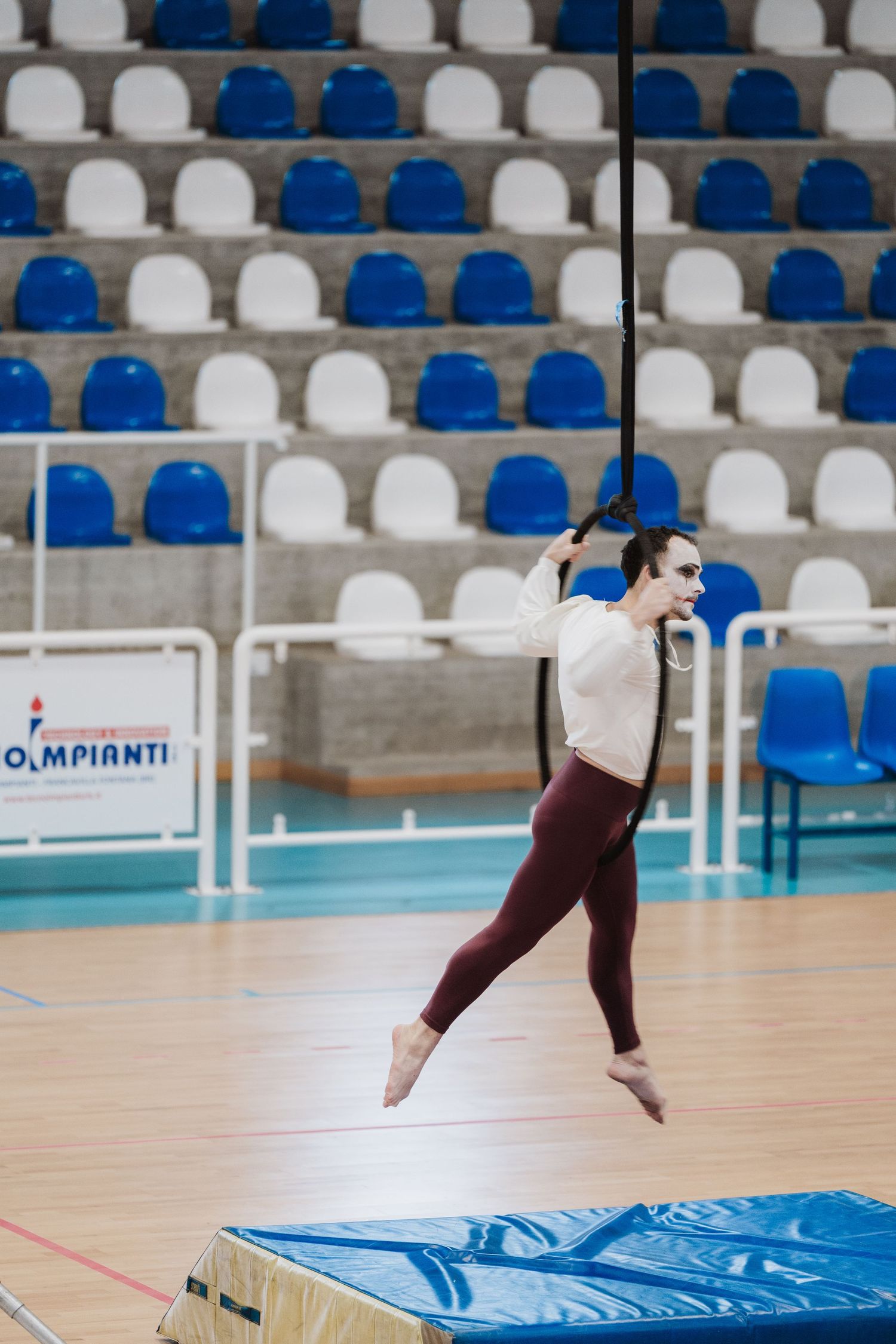 Gymnast in white top and burgundy leggings performs a leap in a sports arena with blue and white stadium seating.
