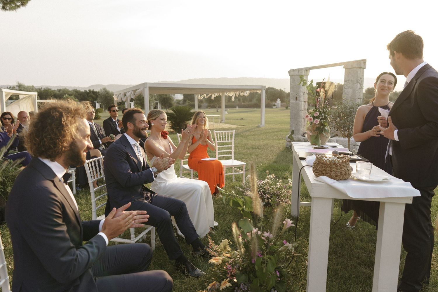 Wedding guests seated outdoors at a ceremony with white chairs and floral arrangements at sunset.
