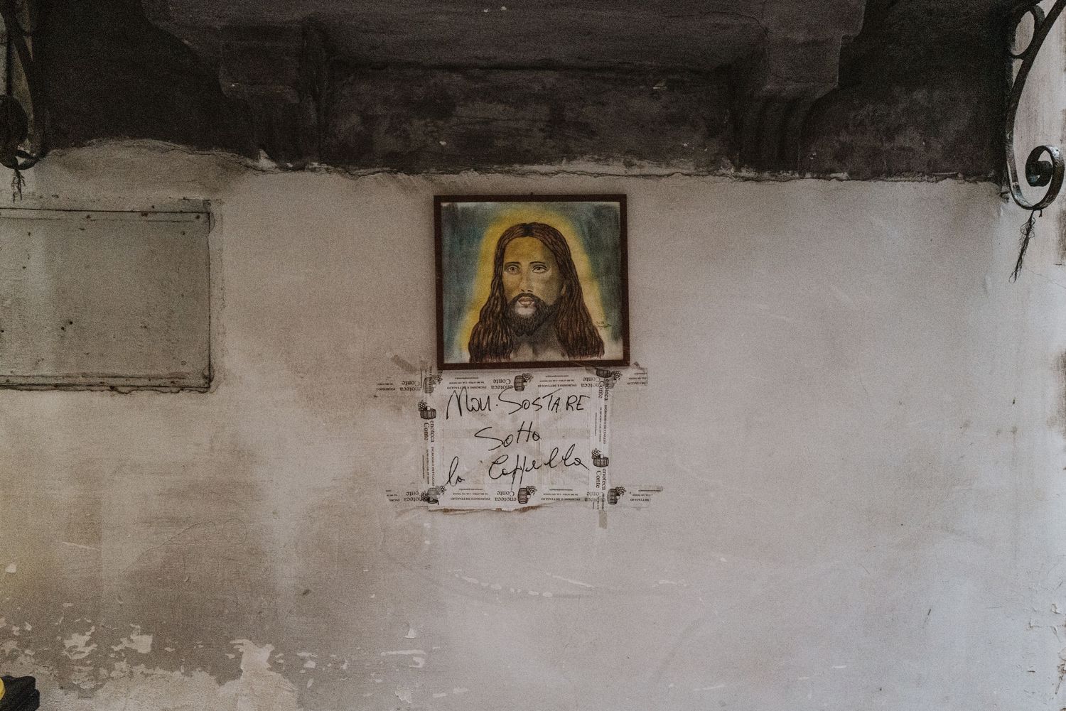A religious portrait hangs on a weathered white wall in a dimly lit space with exposed wooden beams.